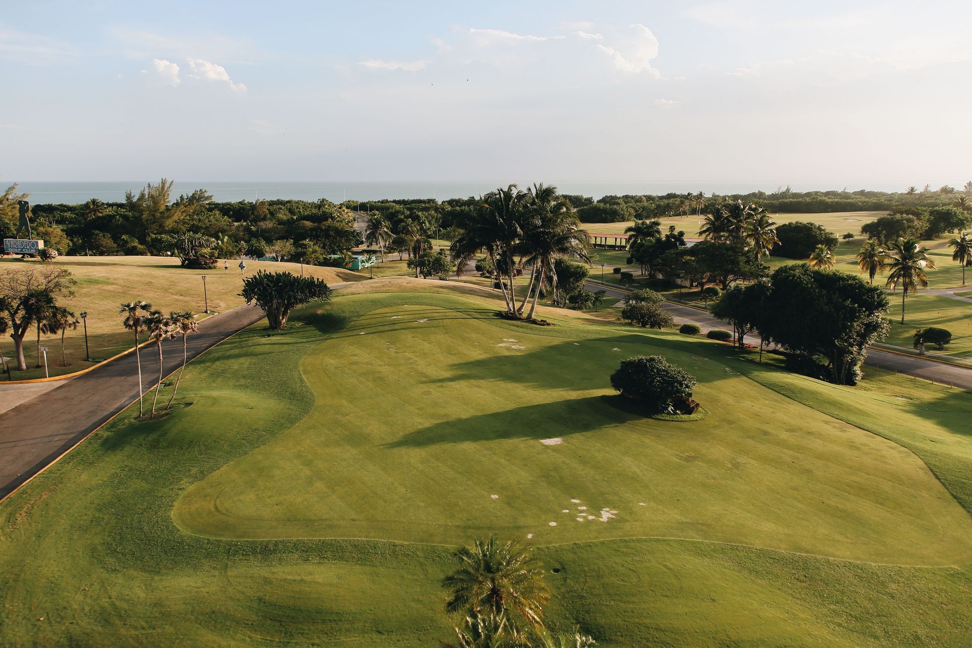 A green golf course fairway with trees, sand traps, and a cart path under a partly cloudy sky