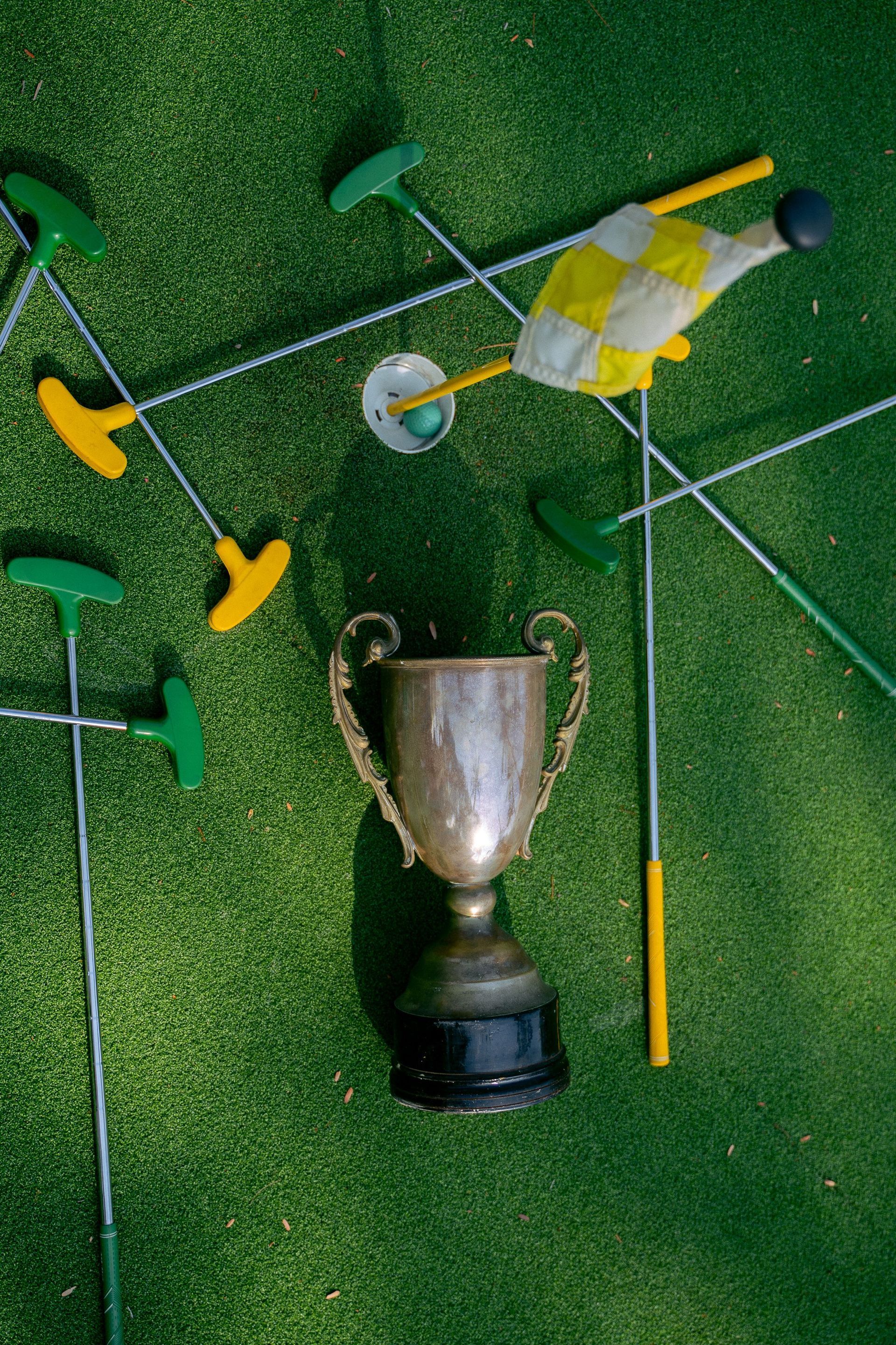 Silver trophy surrounded by green and yellow mini-golf clubs on artificial turf
