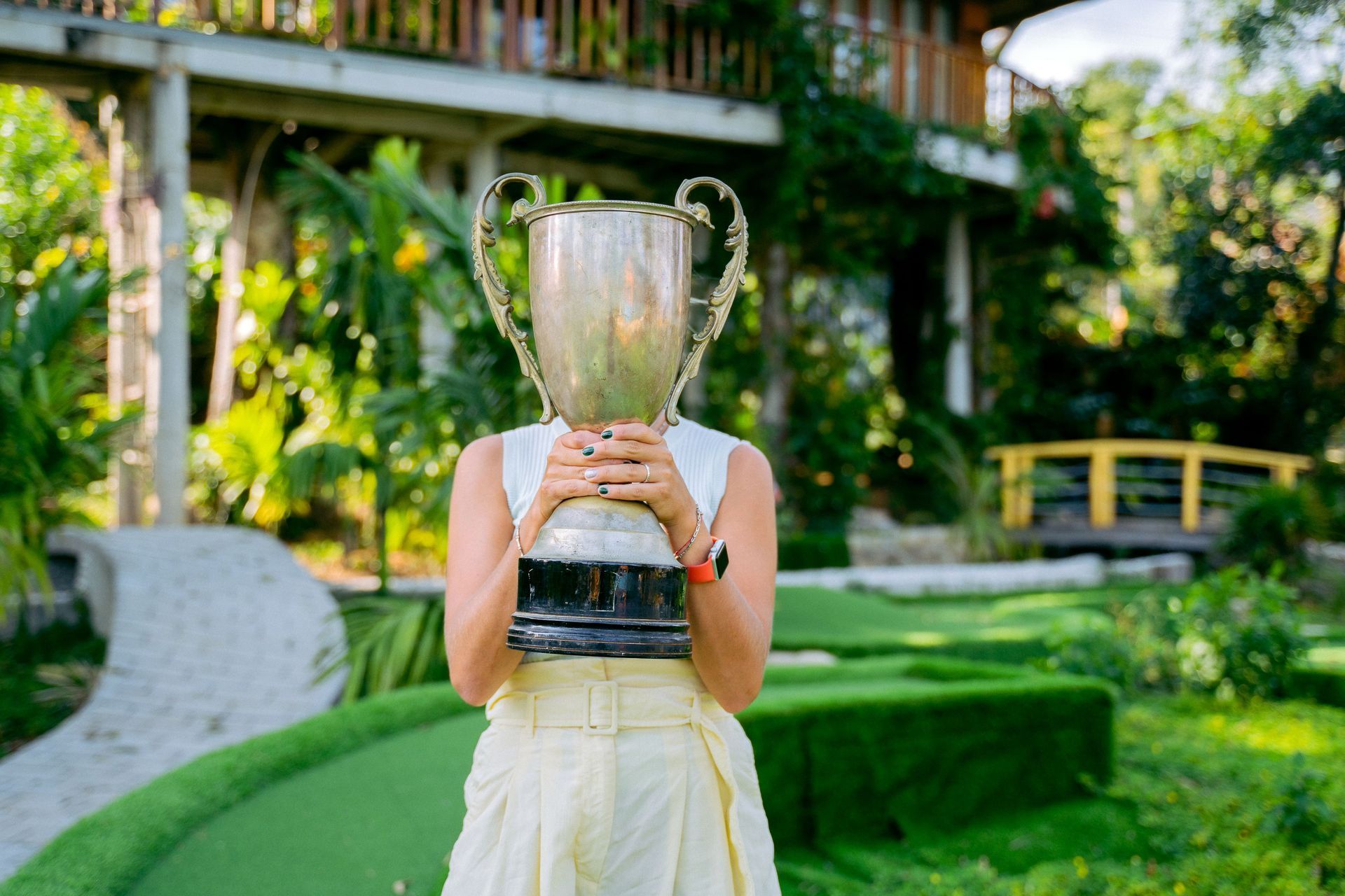 Person holding a large trophy in a lush garden courtyard, with a green bench and walkway in the background