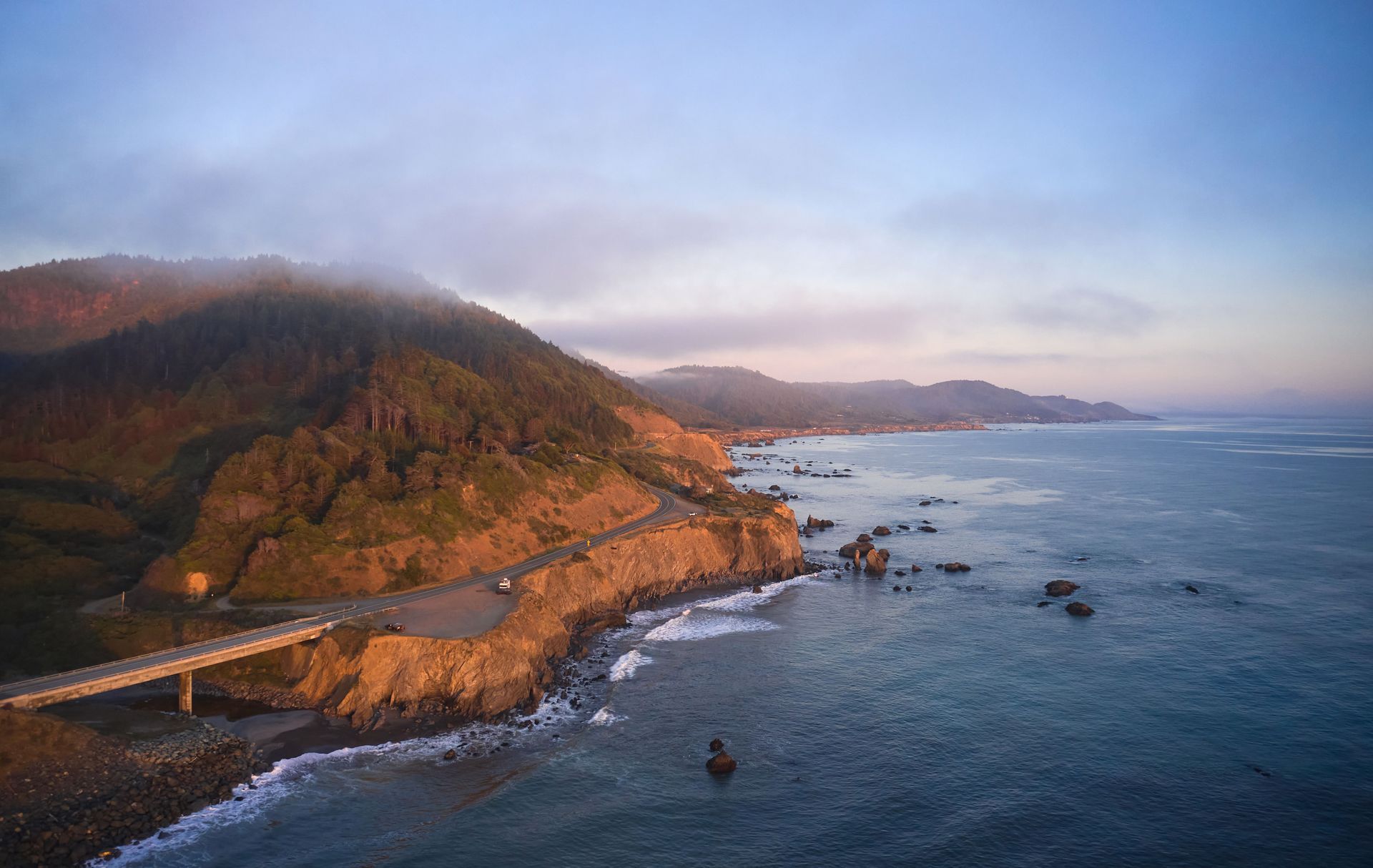 Coastal cliffs at sunset with a bridge, winding road, and calm ocean waves under a cloudy sky