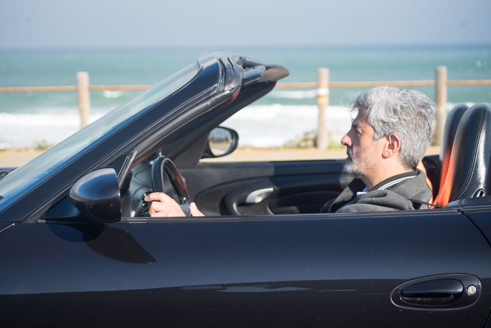 Man driving a black convertible by the seaside, with ocean waves in the background