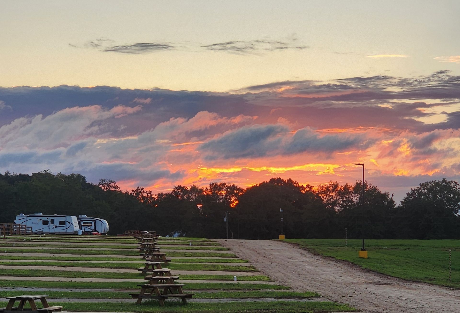 Sunset over a rural campground with RVs, a dirt road, and silhouetted trees beneath pink clouds