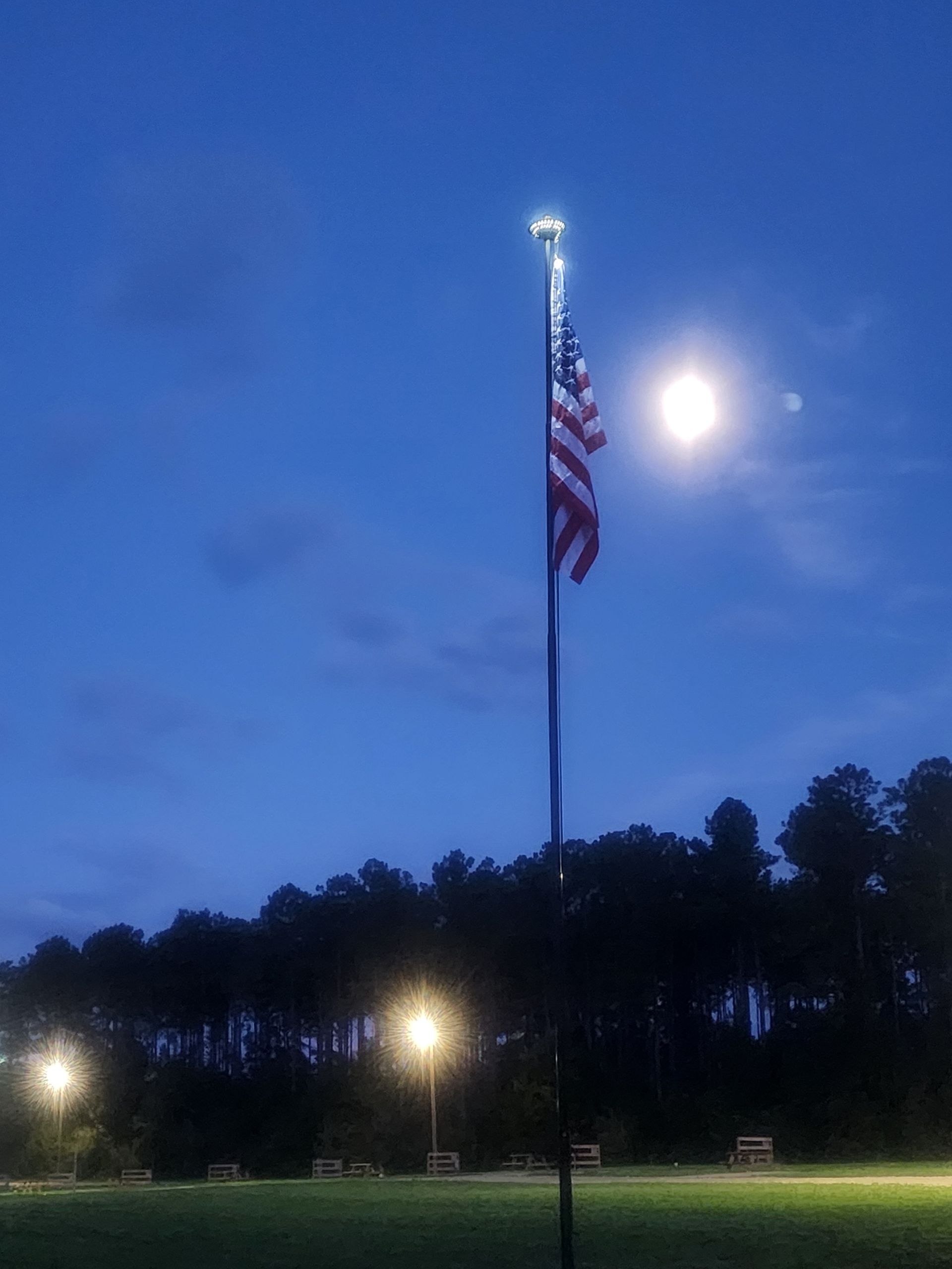Tall flagpole with an American flag at dusk in a lit park under a bright moon.