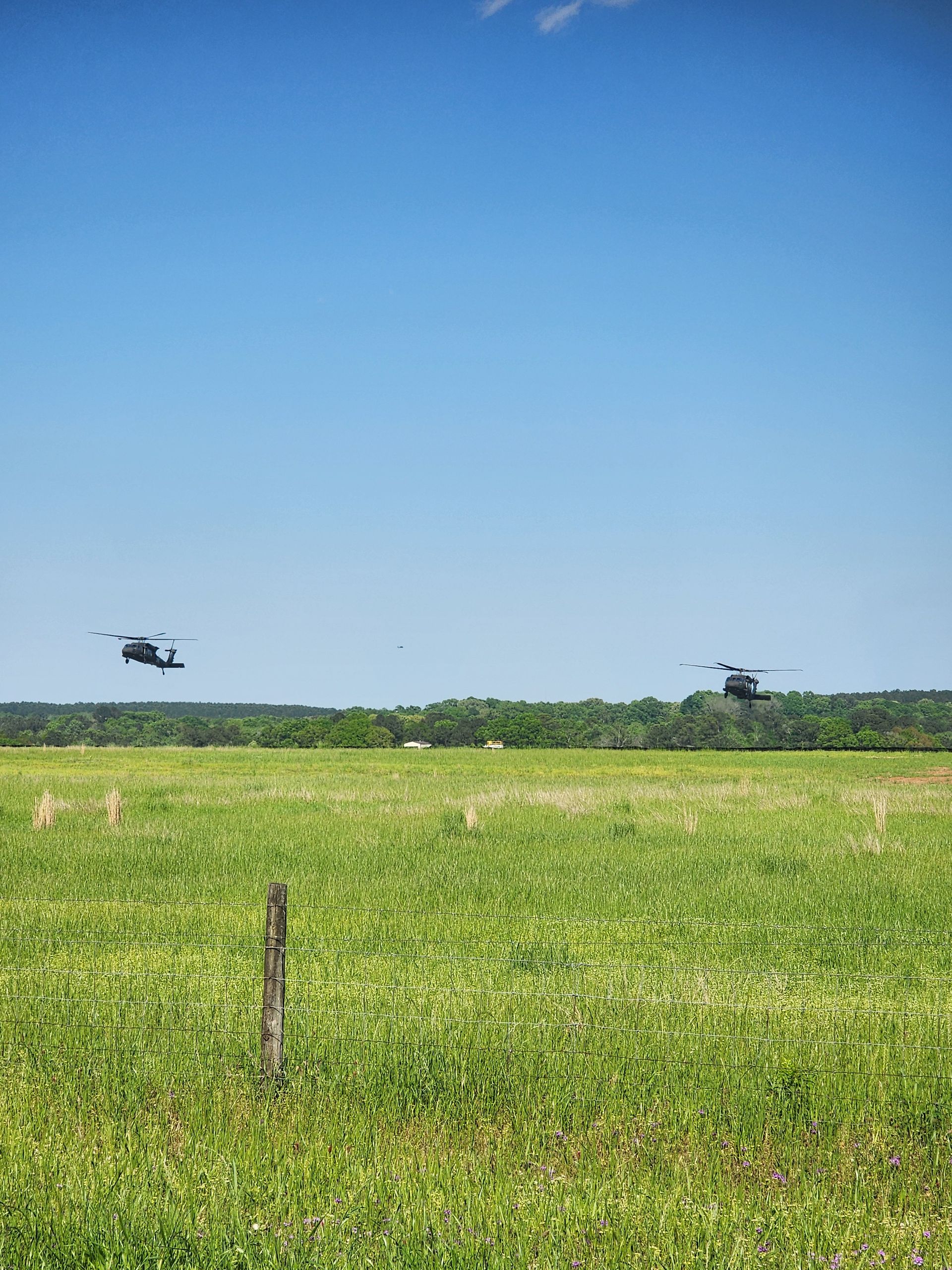 Two helicopters flying over a green field under a clear blue sky