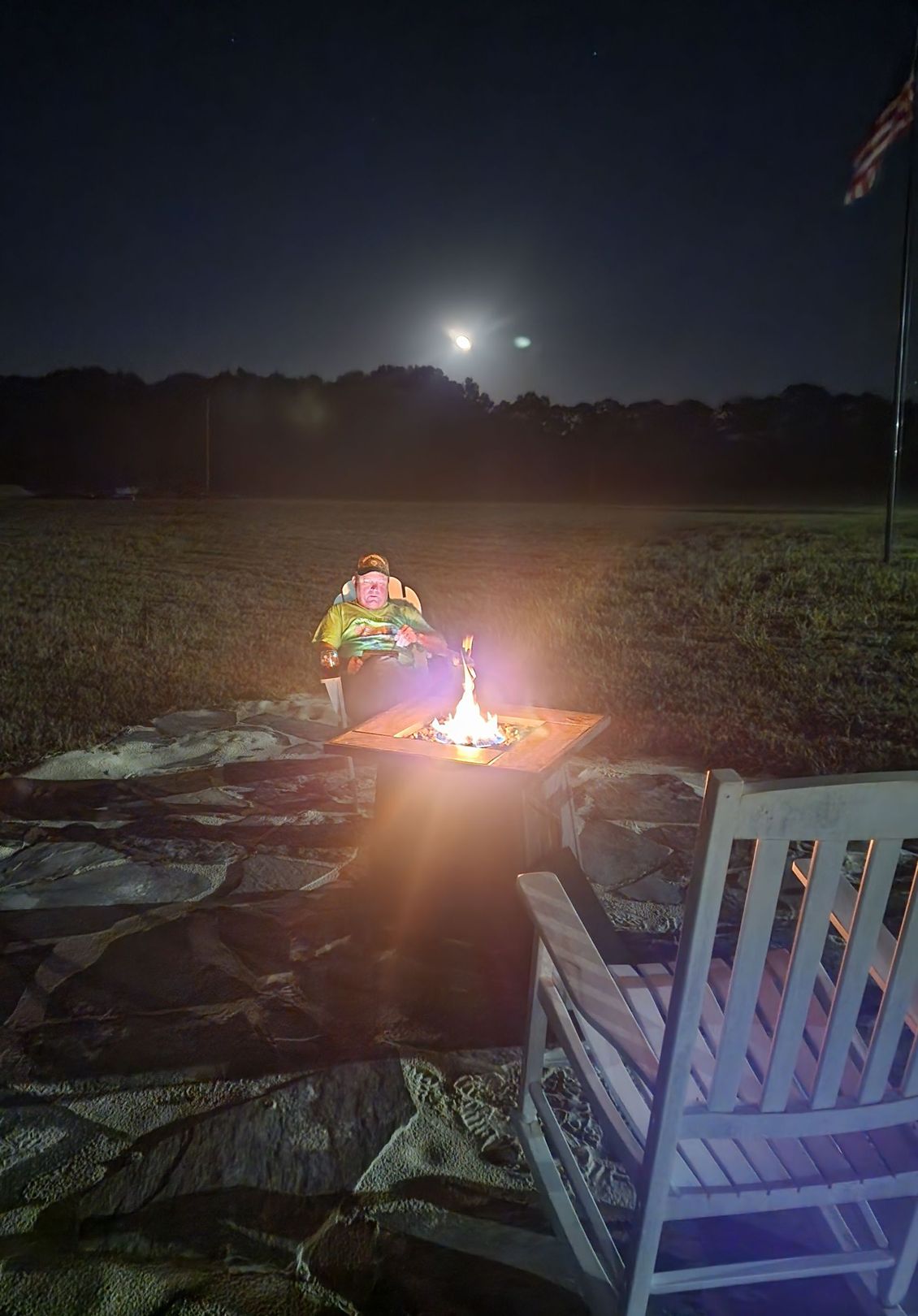 Person sitting by a small campfire at night in a field, with moonlight in the sky