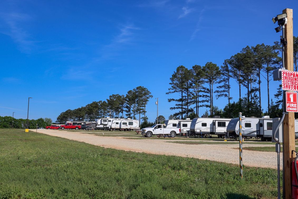 Row of white RVs parked along a gravel lot beside trees under a clear blue sky