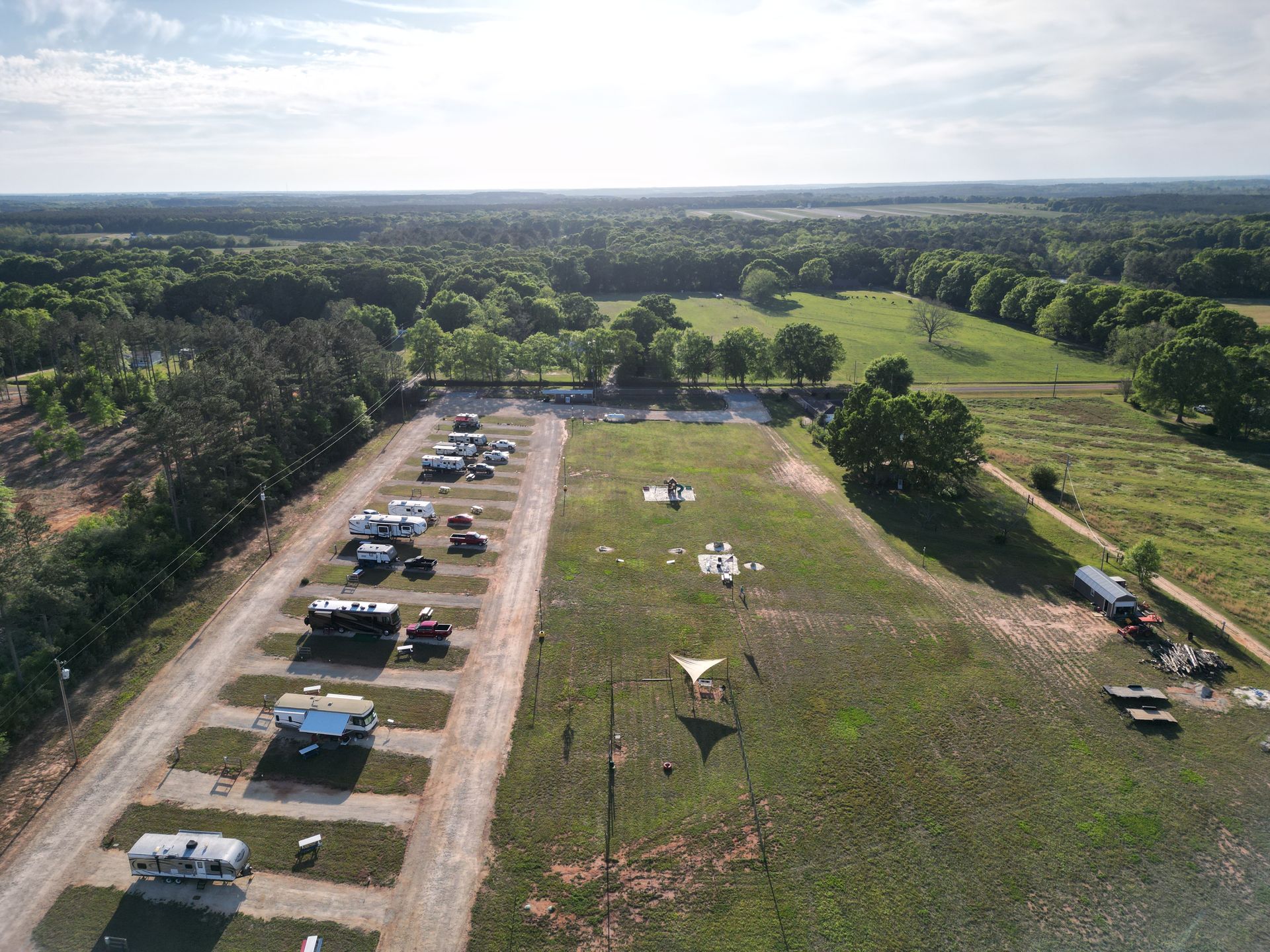Aerial view of a grassy field with parked cars, tents, and trees under a bright sky.