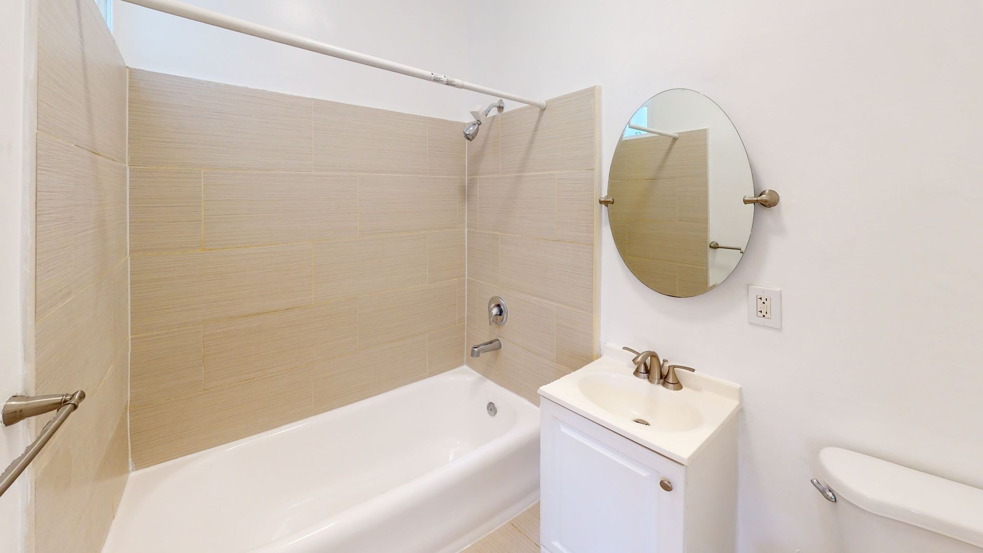 Bathroom with white tub, sink, and toilet; beige tiled shower area, oval mirror.