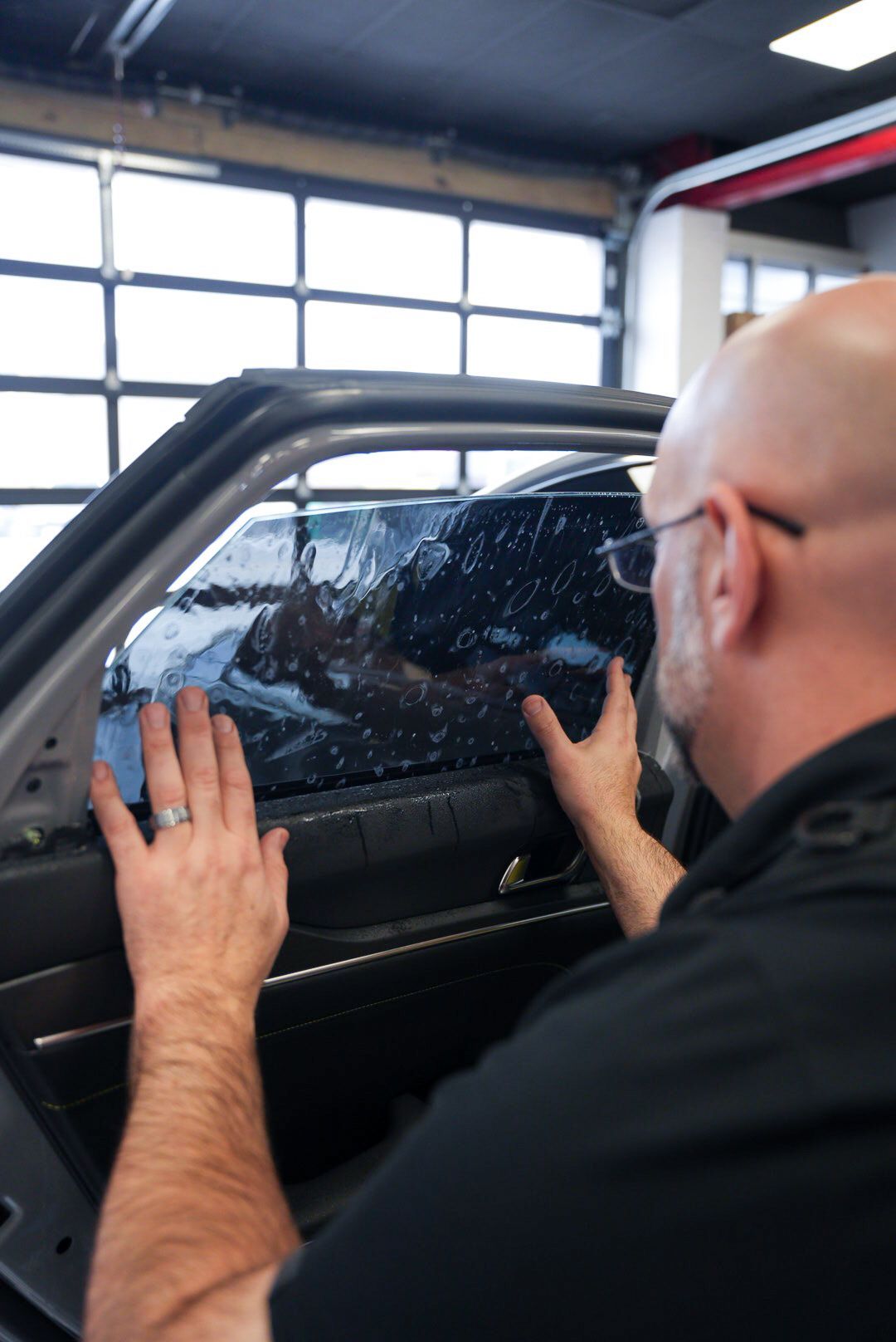 Man applying window tint to a car window in a garage. He's wearing glasses and a black shirt, with his hands working on the dark-tinted glass.
