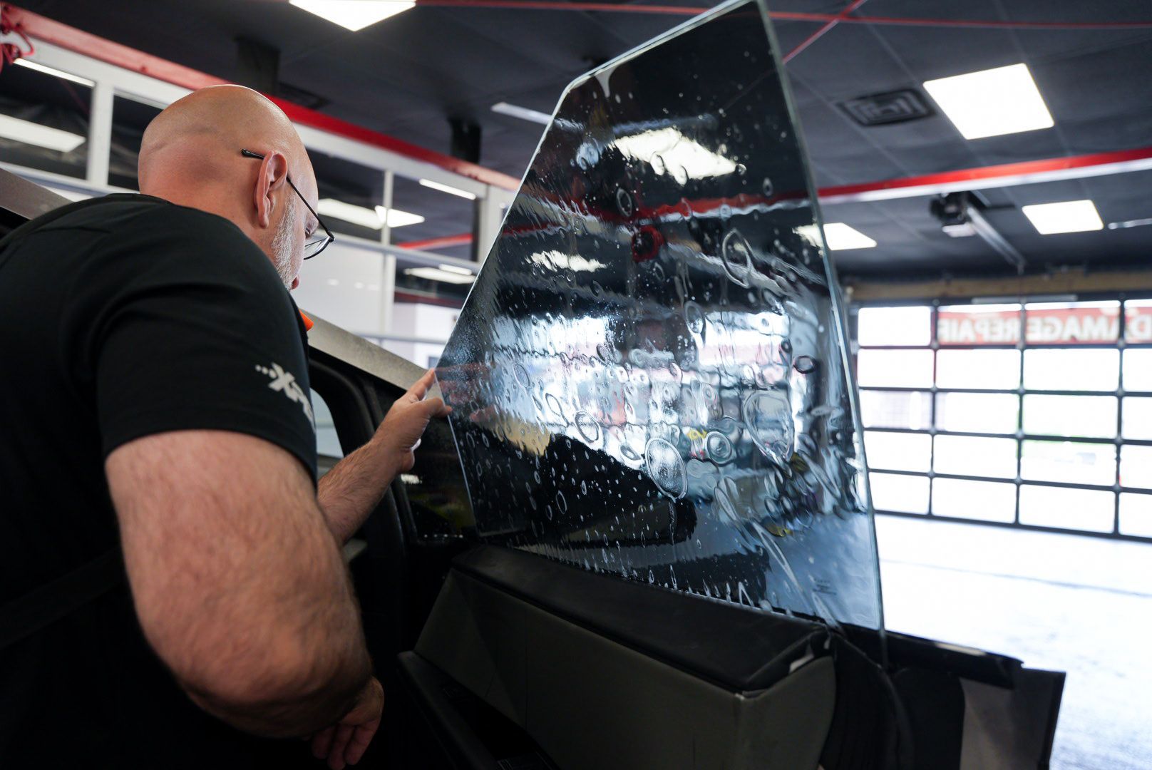 Man applying window tint to a car window in a garage. The window is wet and reflective.