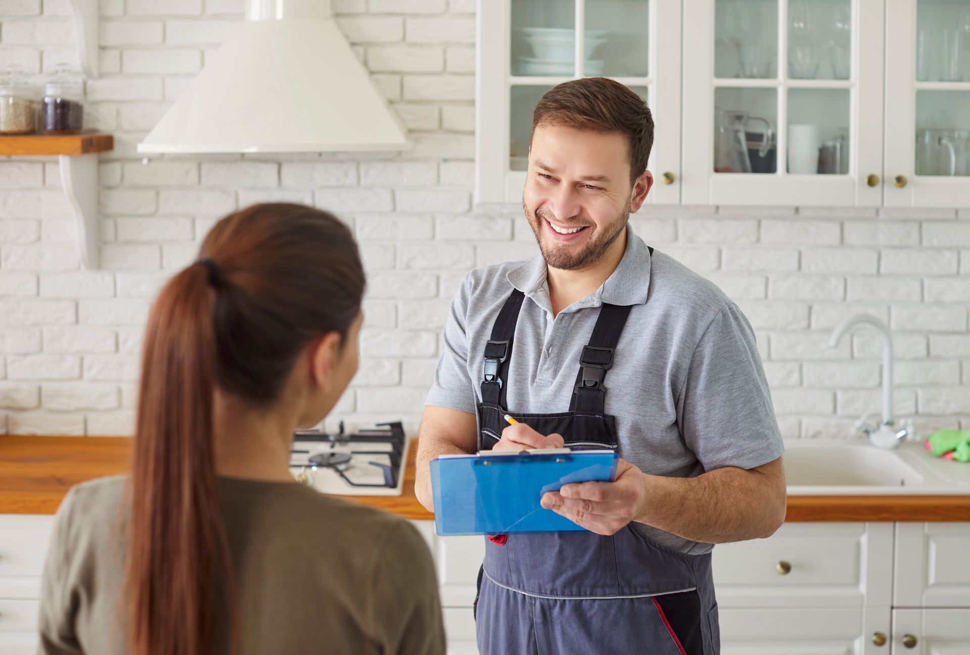 Smiling man in overalls taking notes on a clipboard, talking to a woman in a kitchen.