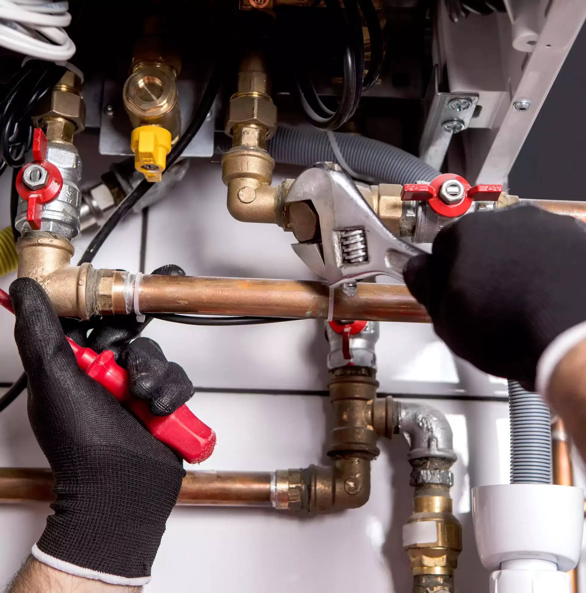 Plumber with black gloves uses a wrench and a tool on copper pipes near a furnace.