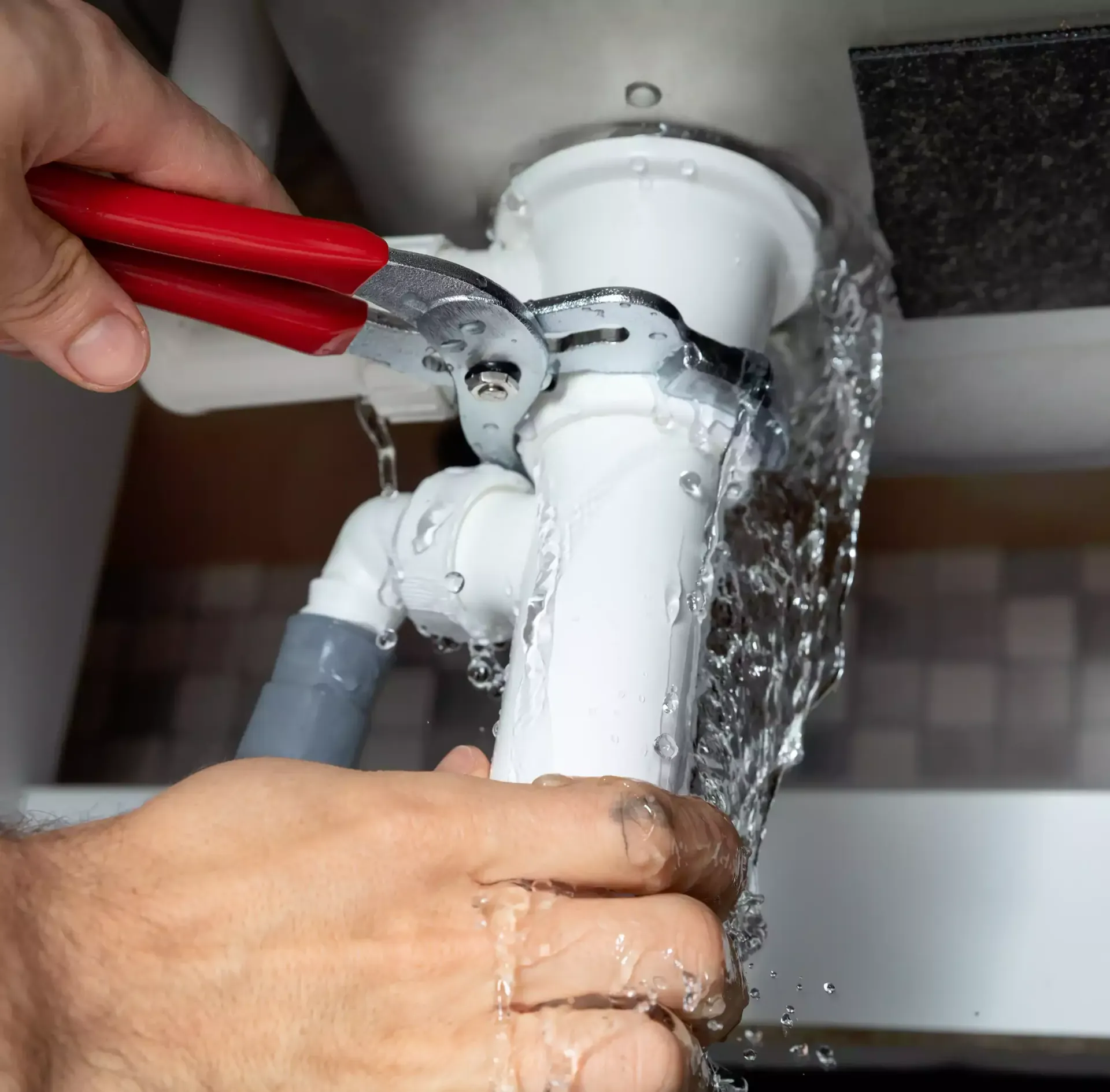 Hands of a person using a wrench on a leaky white drainpipe under a sink; water sprays.
