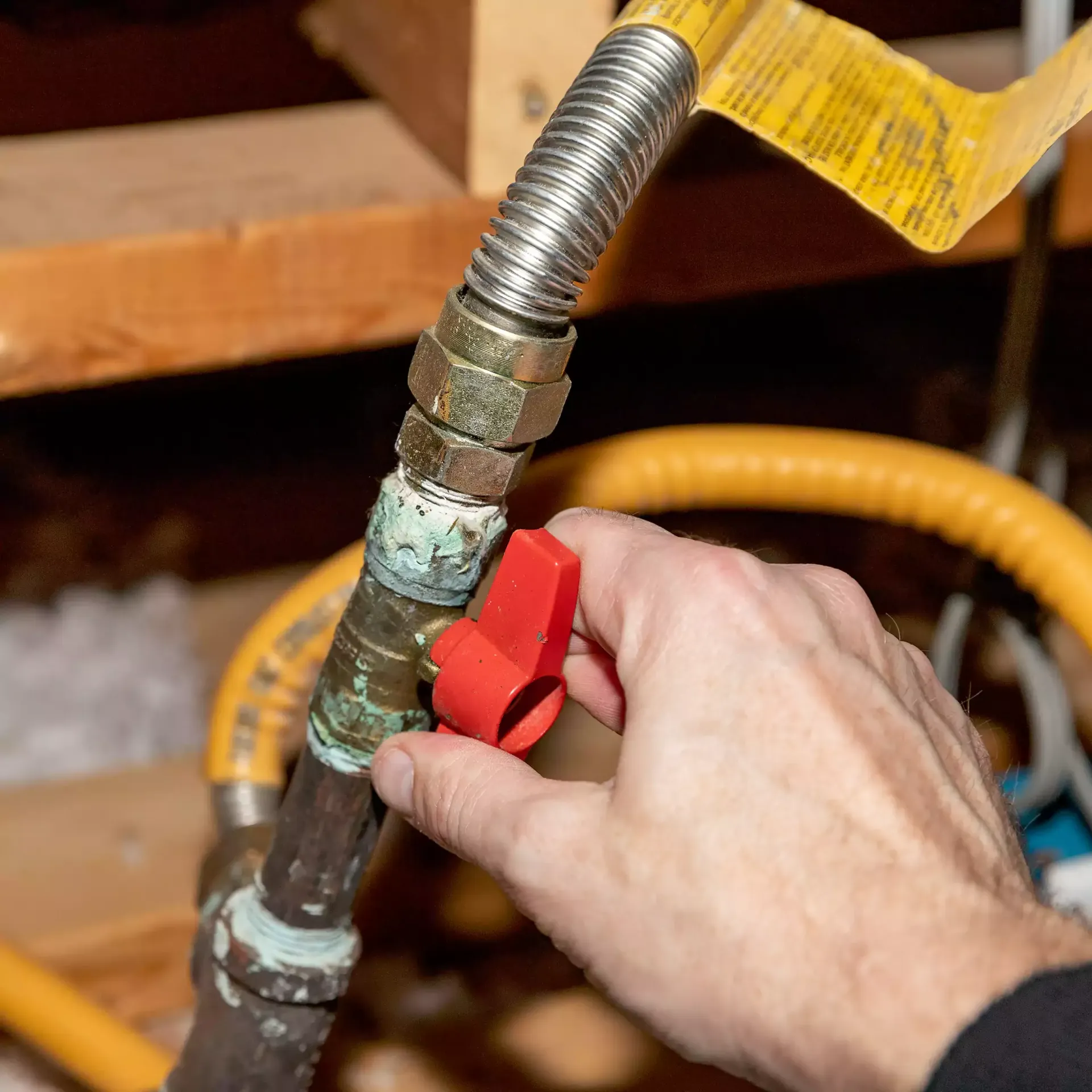 Hand turning a red gas shut-off valve on a copper pipe with a corrugated steel gas line connected.
