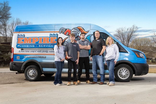 Five people posing in front of a blue Empire Plumbing van with logo and cartoon graphic