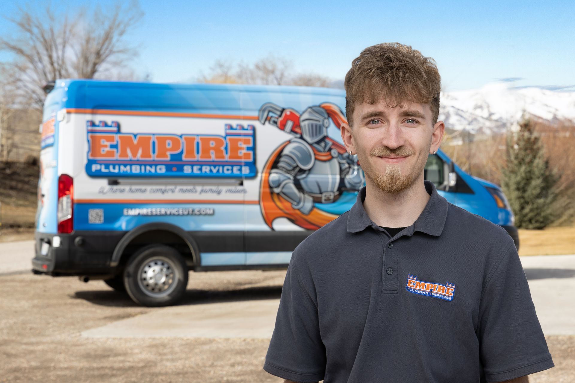 Young man in a navy polo stands before an Empire Plumbing van with a knight logo outdoors.
