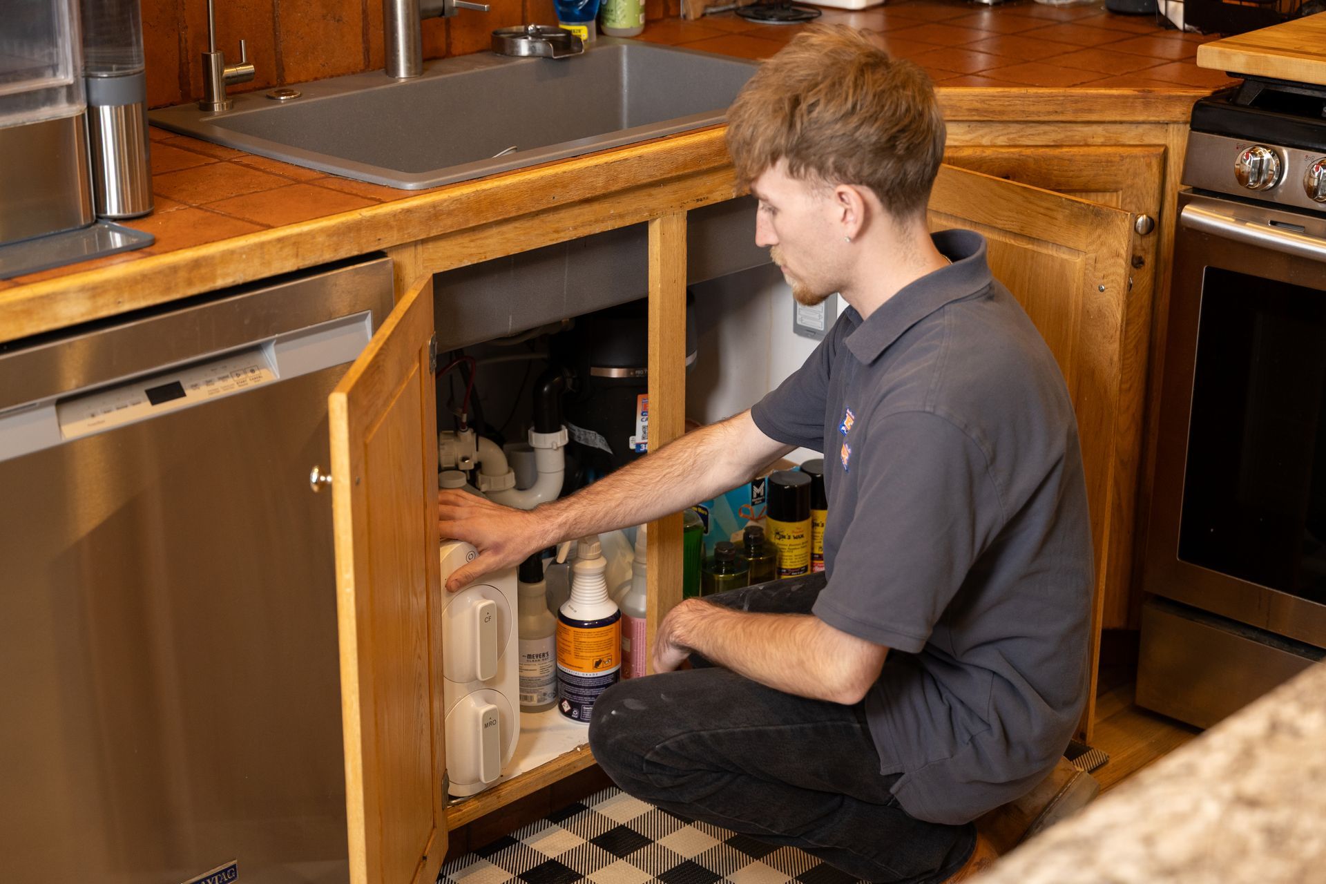 Person kneeling under kitchen sink, reaching into open cabinet beside dishwasher and stove.