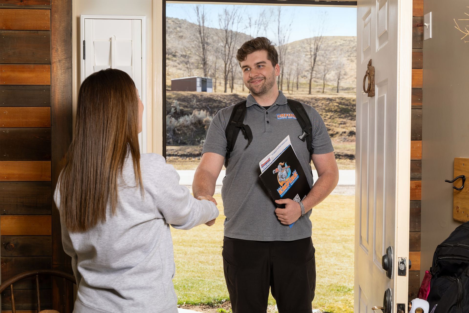 Two people greet at an open doorway, one holding books and a backpack, with a sunny yard outside.