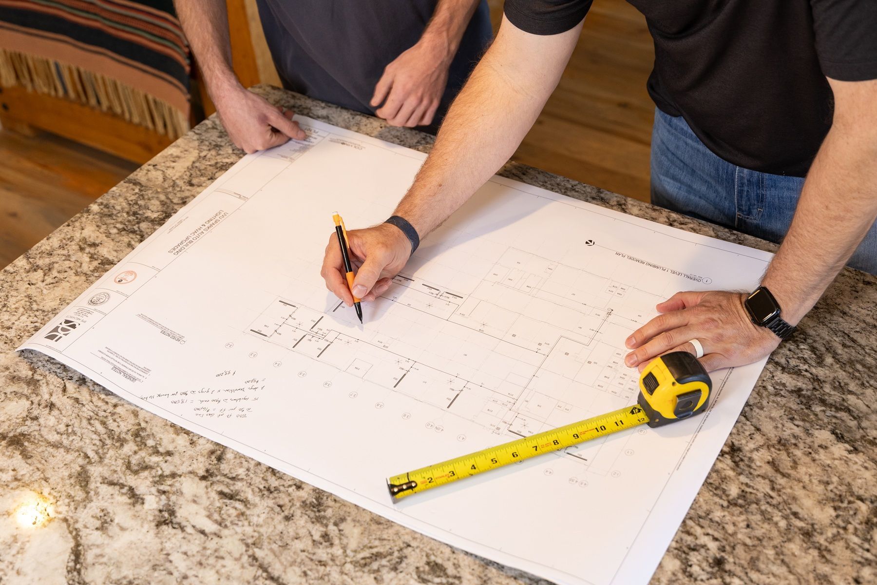 Two people marking measurements on a large white floor plan with a pencil and tape measure on a countertop Two people marking measurements on a large white floor plan with a pencil and tape measure on a countertop