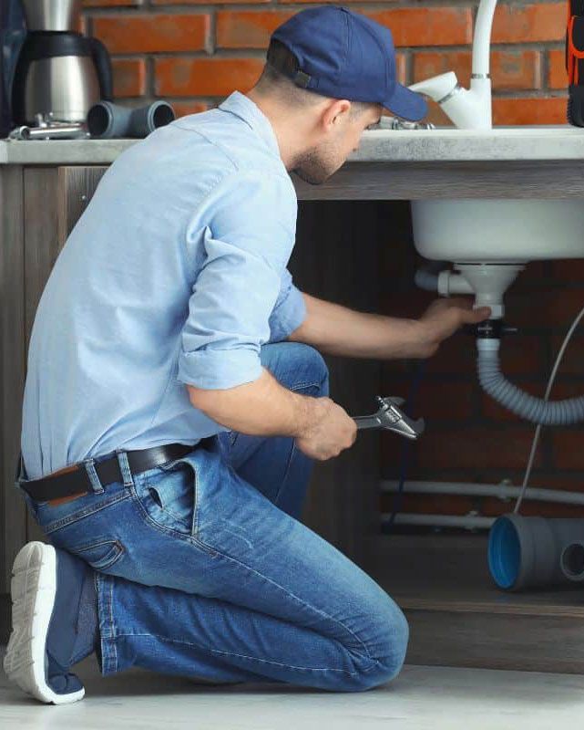 Plumber kneeling under a sink, using a wrench to fix the plumbing.