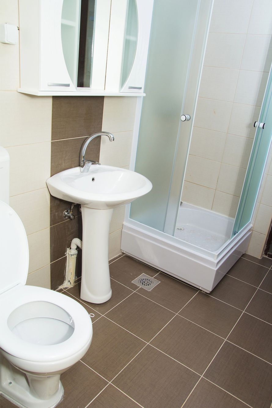 Small bathroom with toilet, pedestal sink, and shower stall on brown tiled floor.