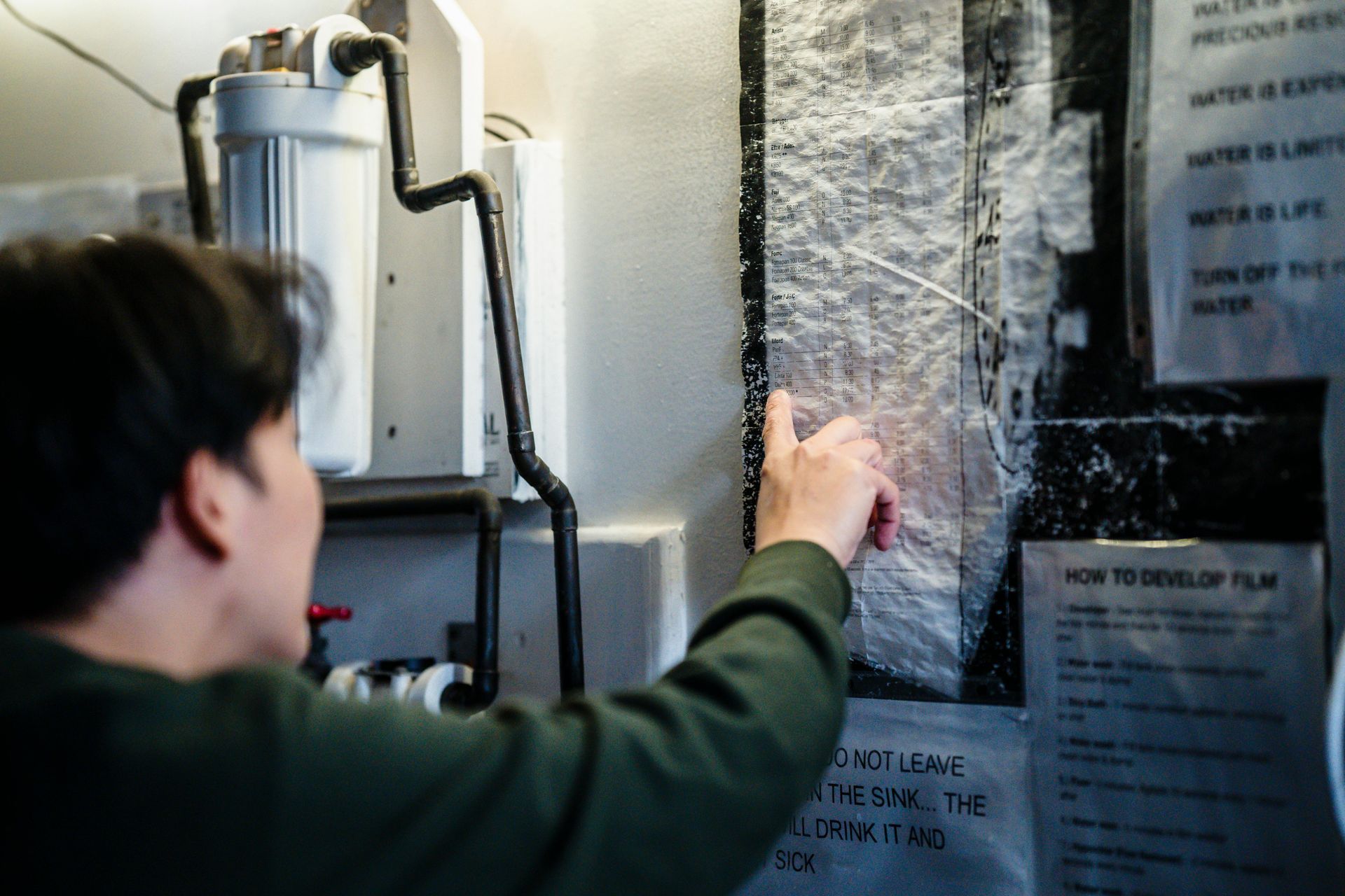 Person points at film development instructions on a wall. A water filter and pipes are visible.