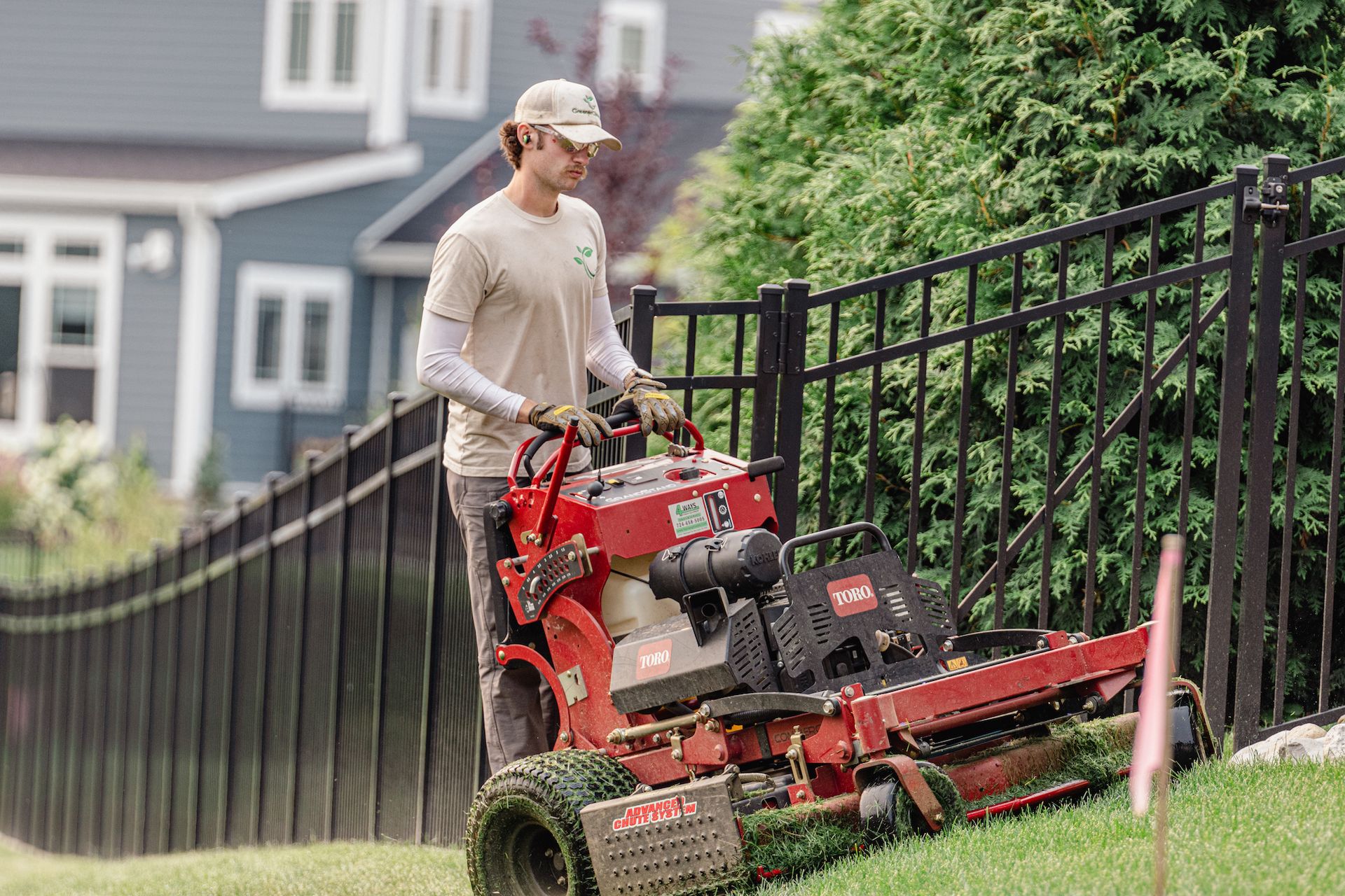 Person mowing a grassy slope with a red riding mower beside a black fence