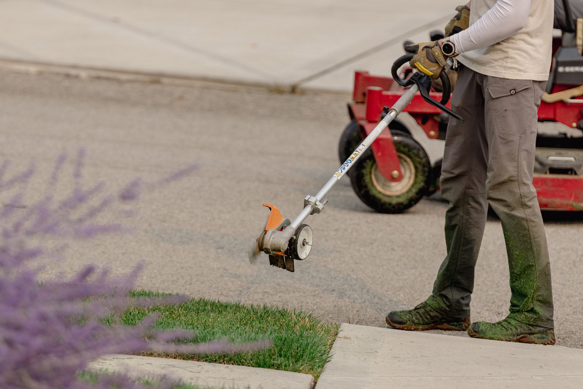Person using a string trimmer to cut grass along a sidewalk near a red lawn mower