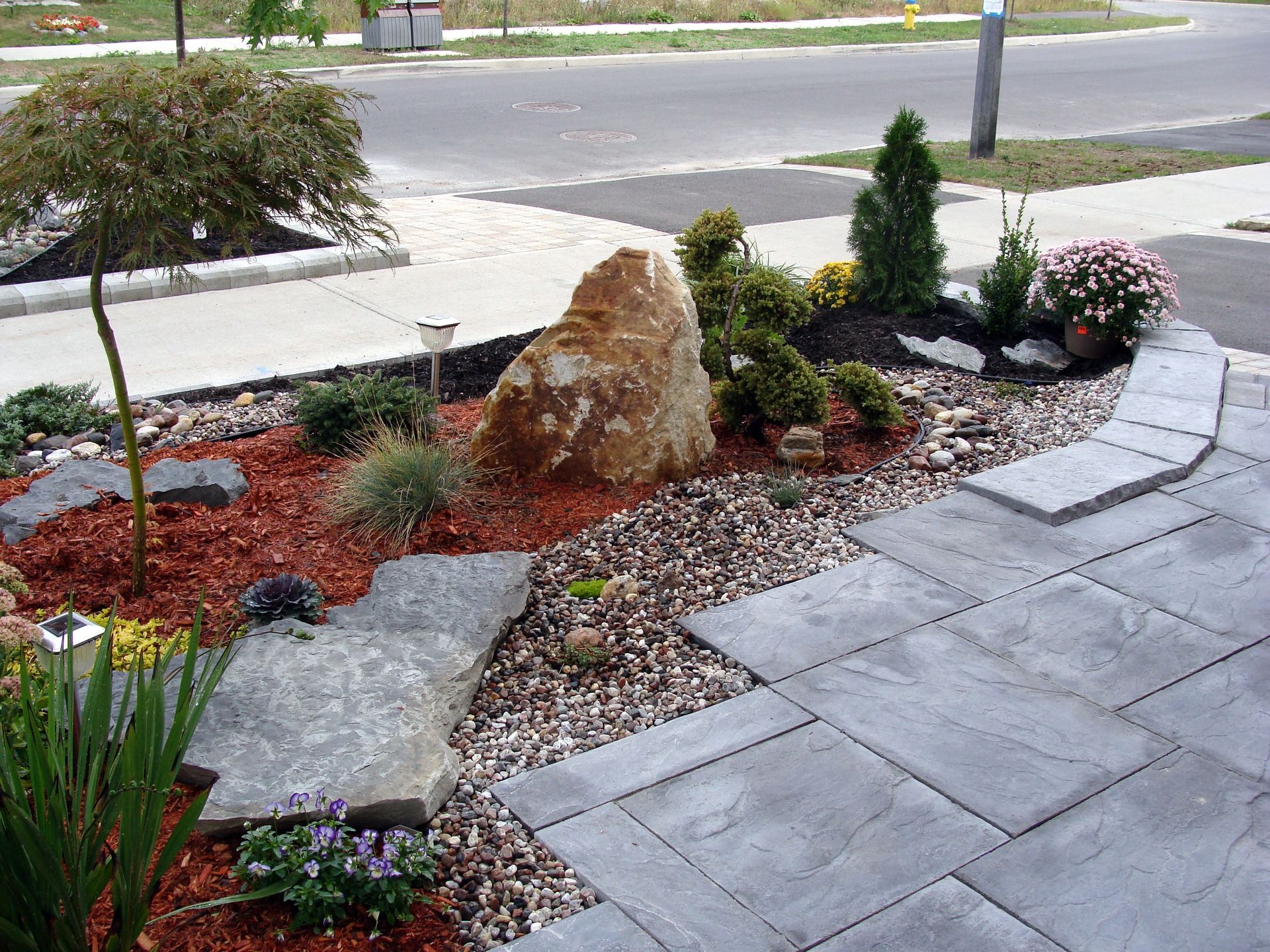 Landscaped front yard with boulders, shrubs, red mulch, and a paved walkway beside the street