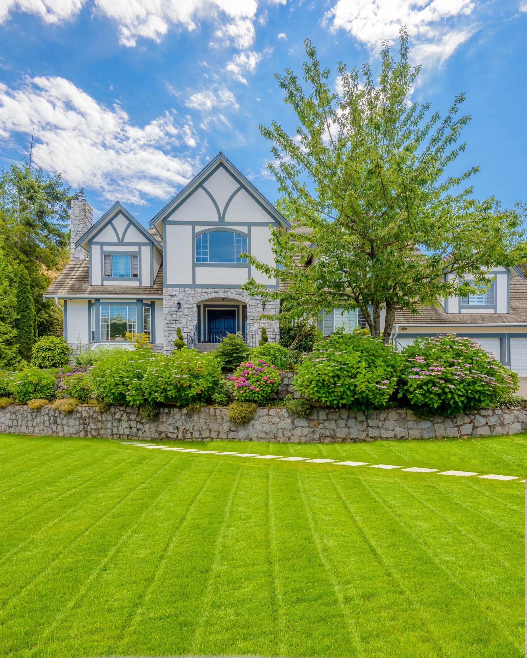 Large suburban house with gray trim, green lawn, and landscaped front yard under a blue sky