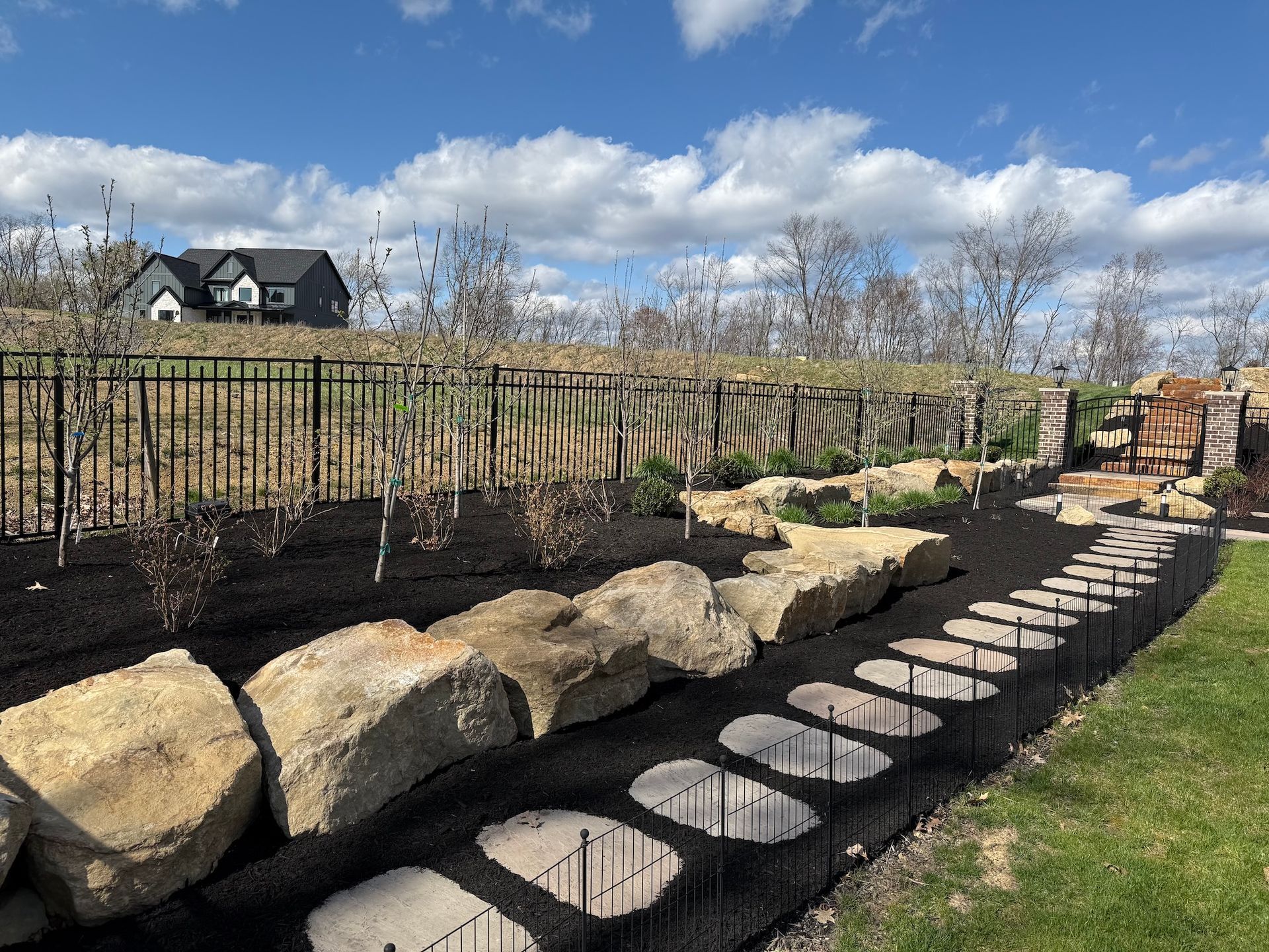 Stone steps beside a landscaped garden wall with large rocks, grass, and a metal fence under a cloudy sky