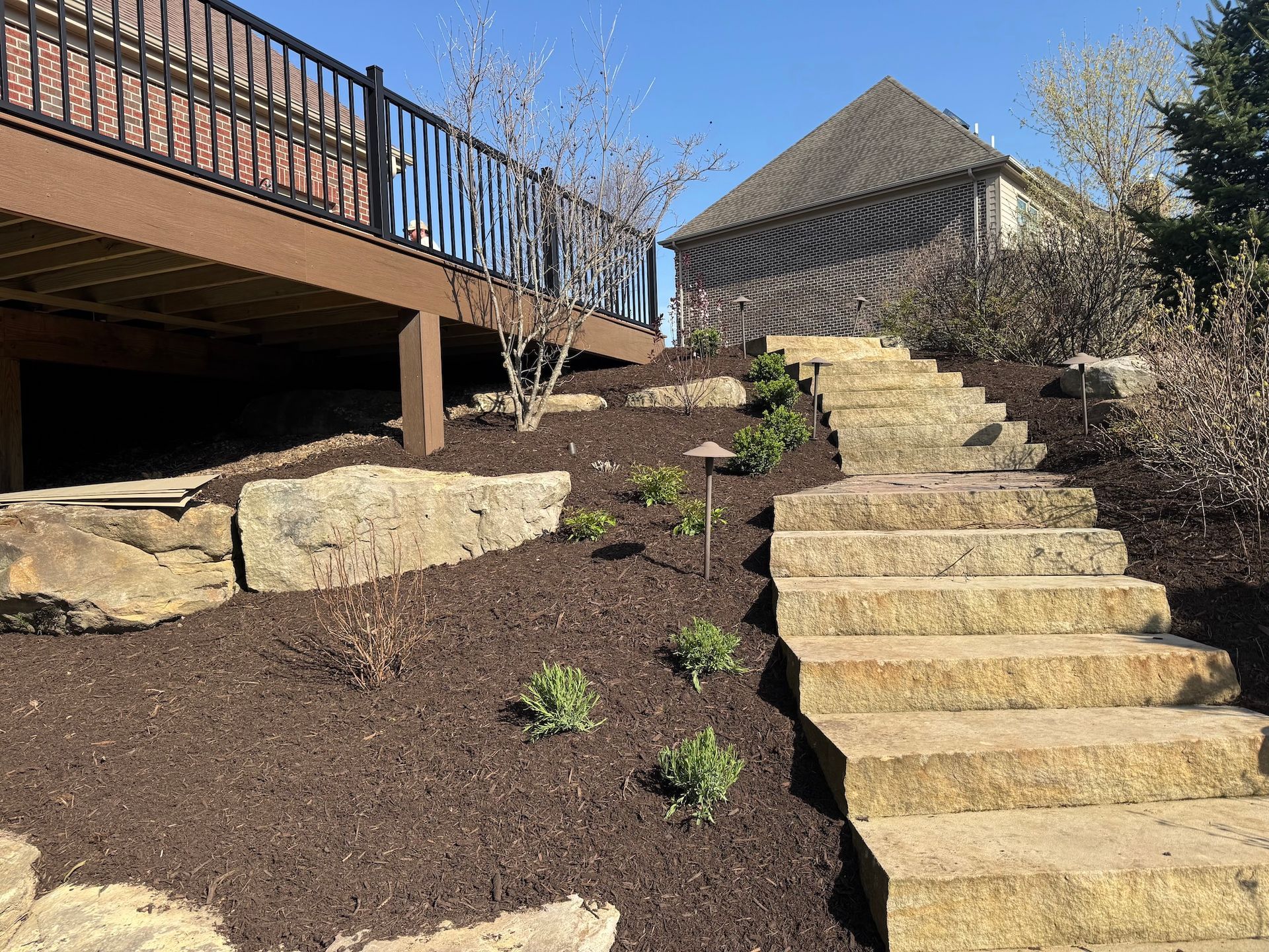 Stone steps beside a mulched hillside leading to a deck and a small shed in a sunny yard