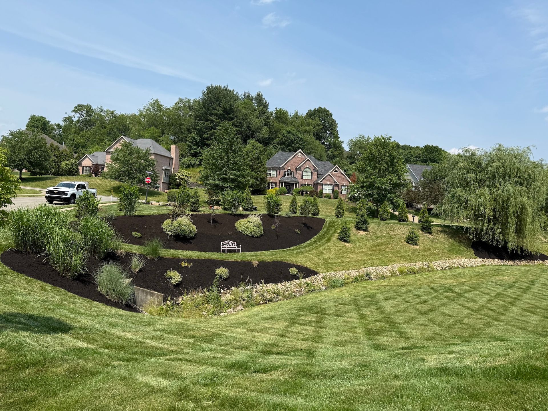 Landscaped hillside garden with dark mulch beds, shrubs, and houses beyond a mowed lawn under a blue sky