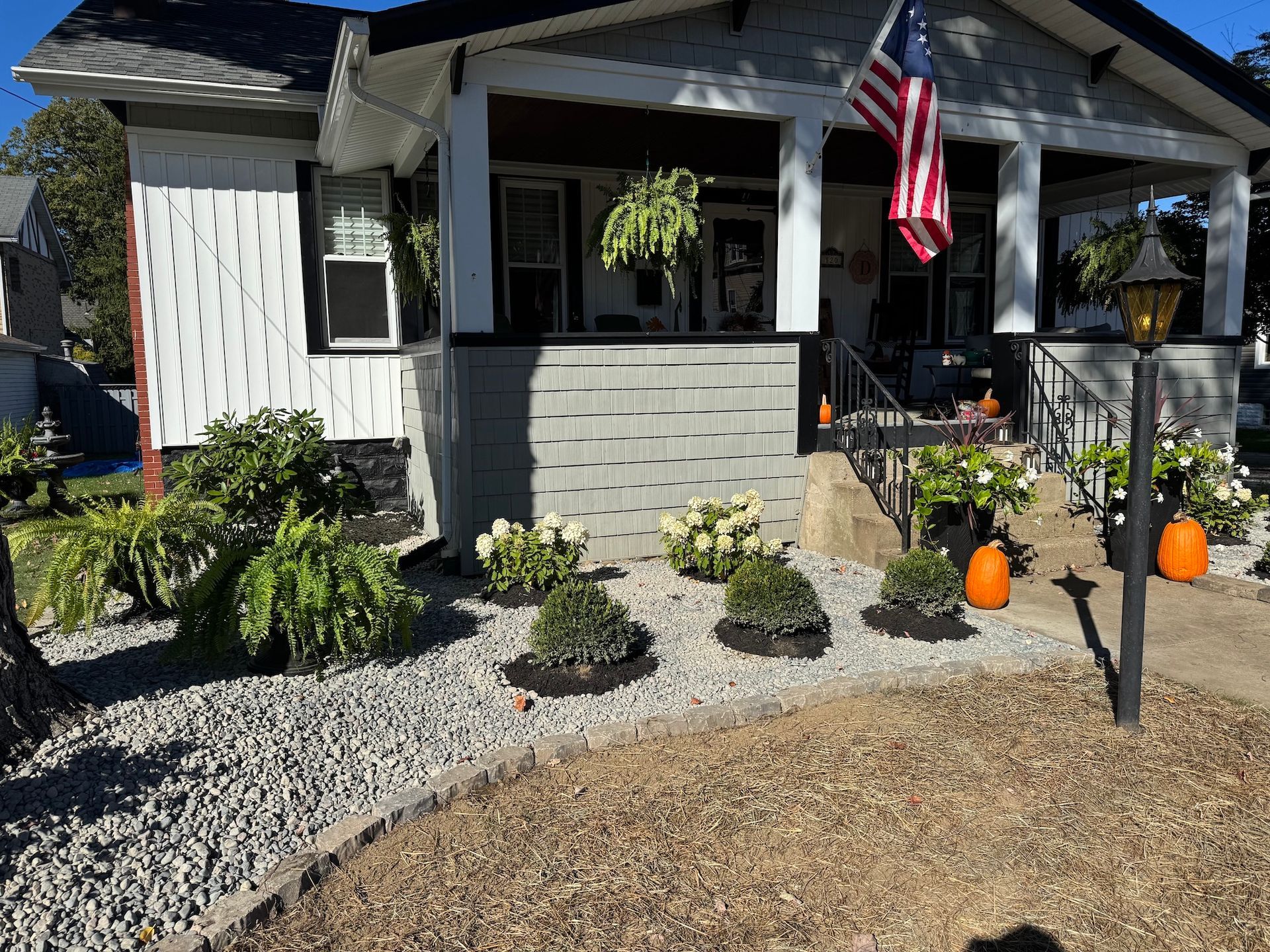 Front porch with American flag, hanging fern, pumpkins, and gravel landscaping in front of a gray house