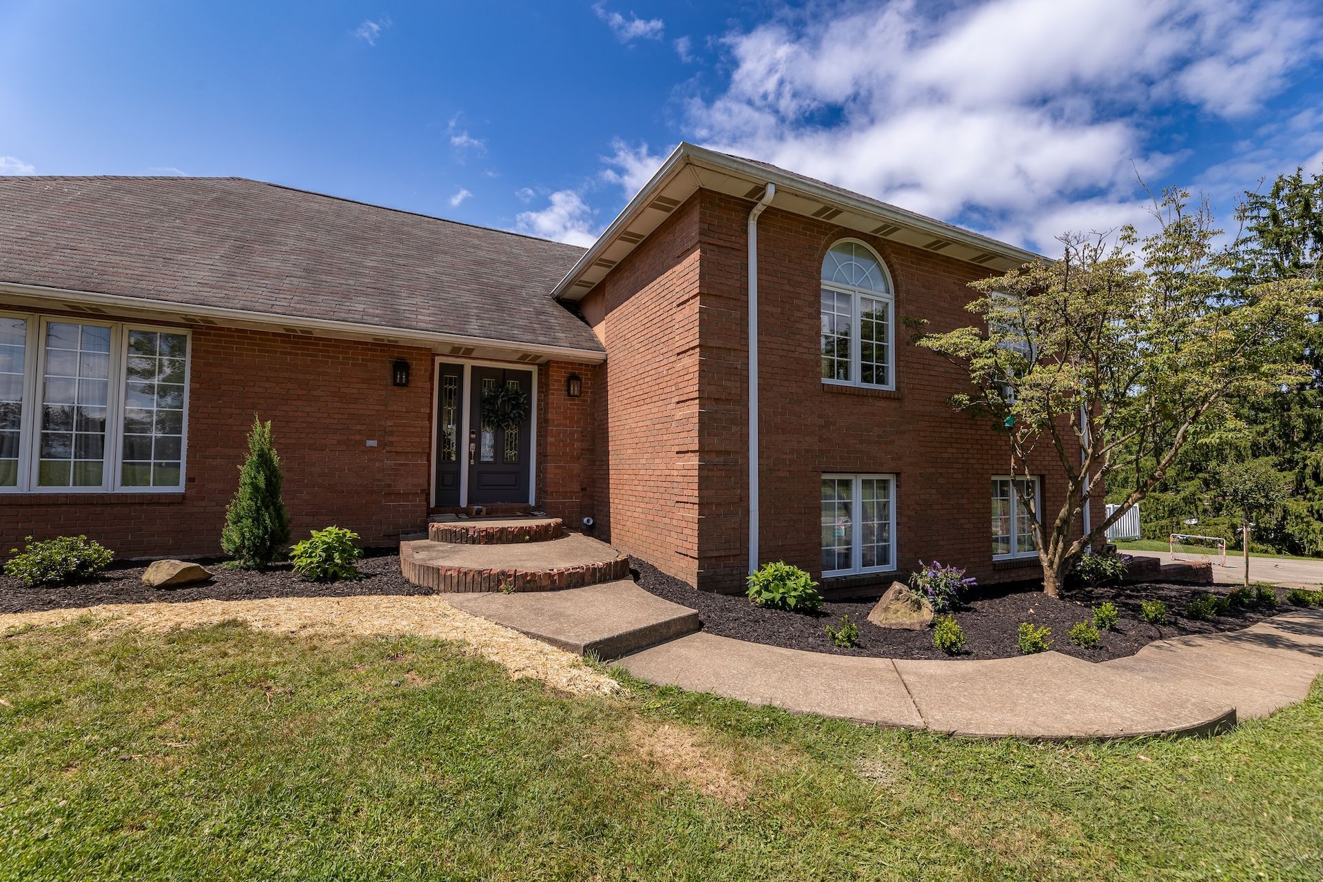 Brick house exterior with front steps, curved walkway, and green lawn under a partly cloudy sky