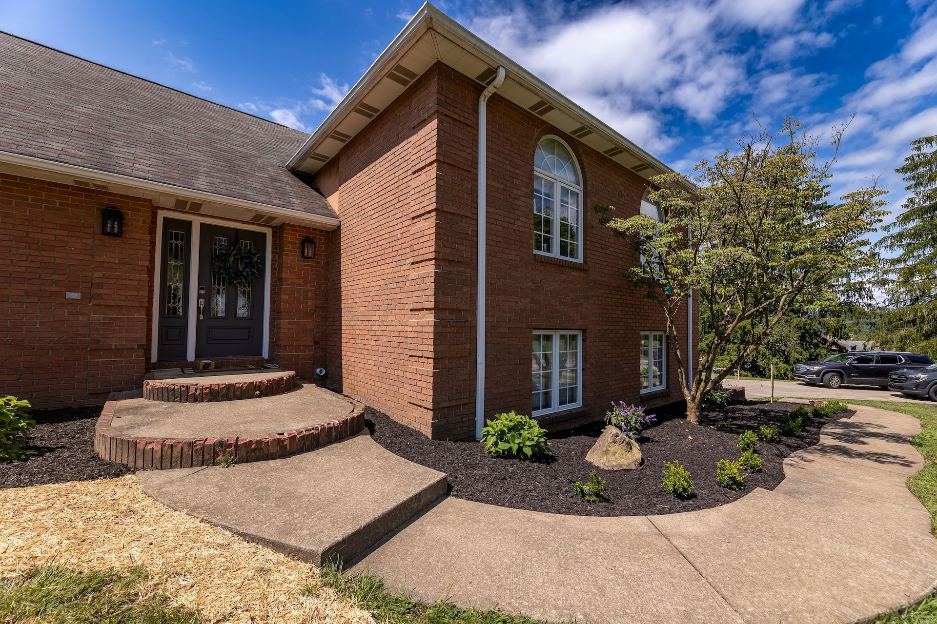 Brick house entrance with curved concrete walkway, steps, and landscaped front yard under a blue sky