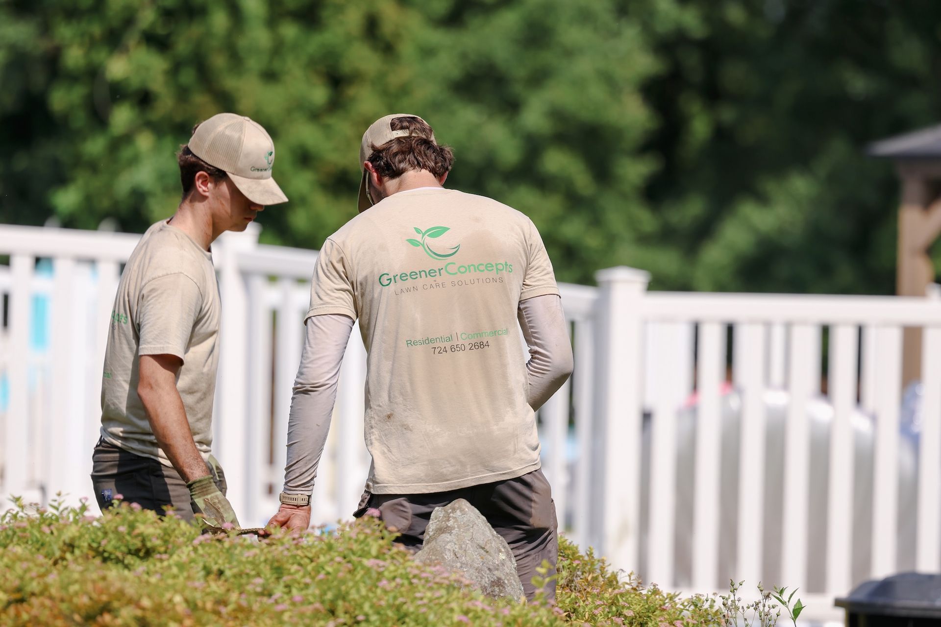 Two workers in tan shirts stand by a white fence in a sunny yard.