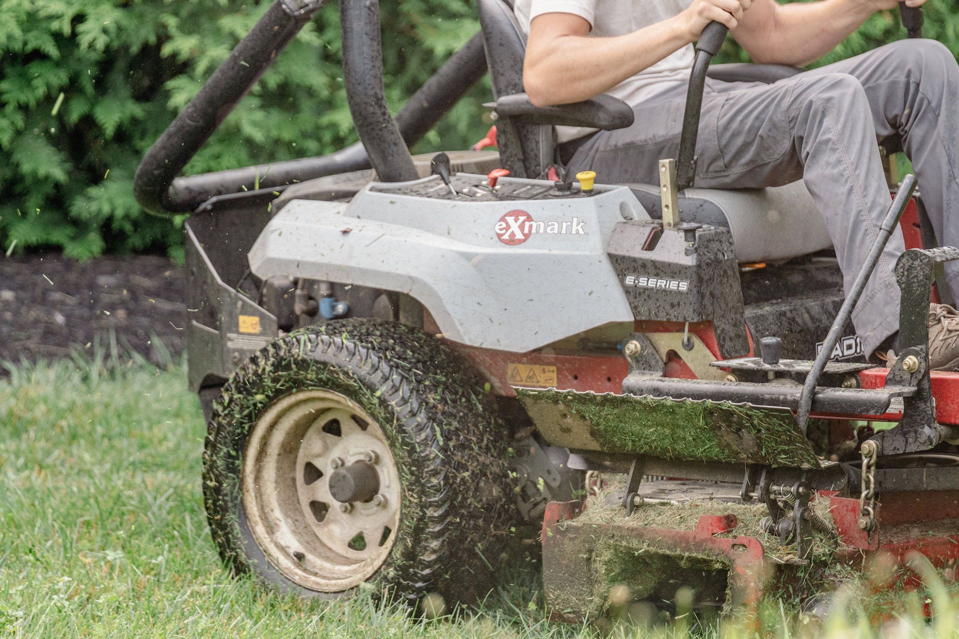 A person drives a gray off-road mower through grass, with a large rear tire in view.