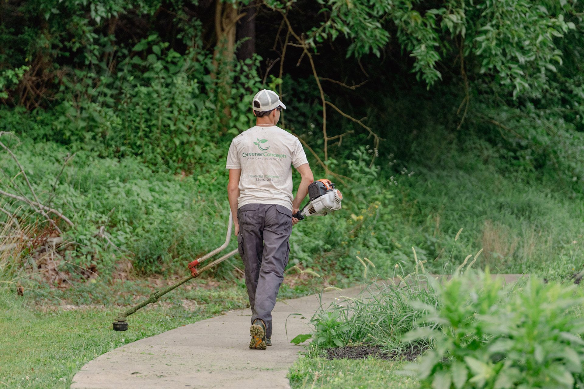 Person using a string trimmer along a concrete path beside dense green vegetation