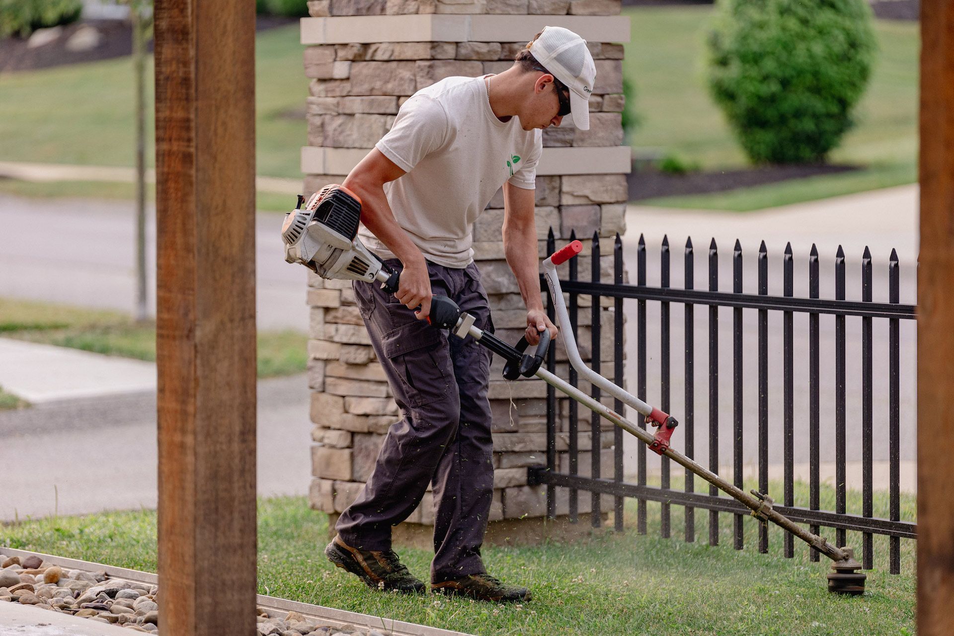 Worker trimming grass along a black fence with a gas-powered string trimmer.