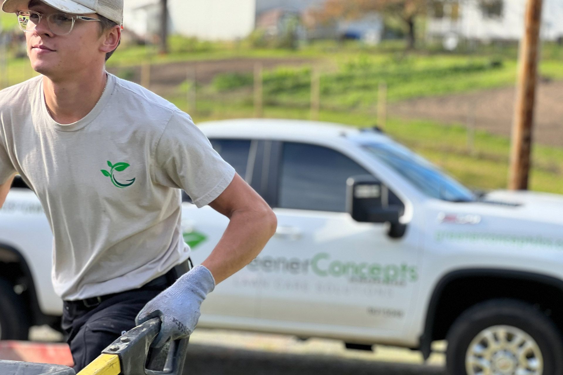Worker in a gray T-shirt and cap standing beside a white pickup truck on a grassy roadside