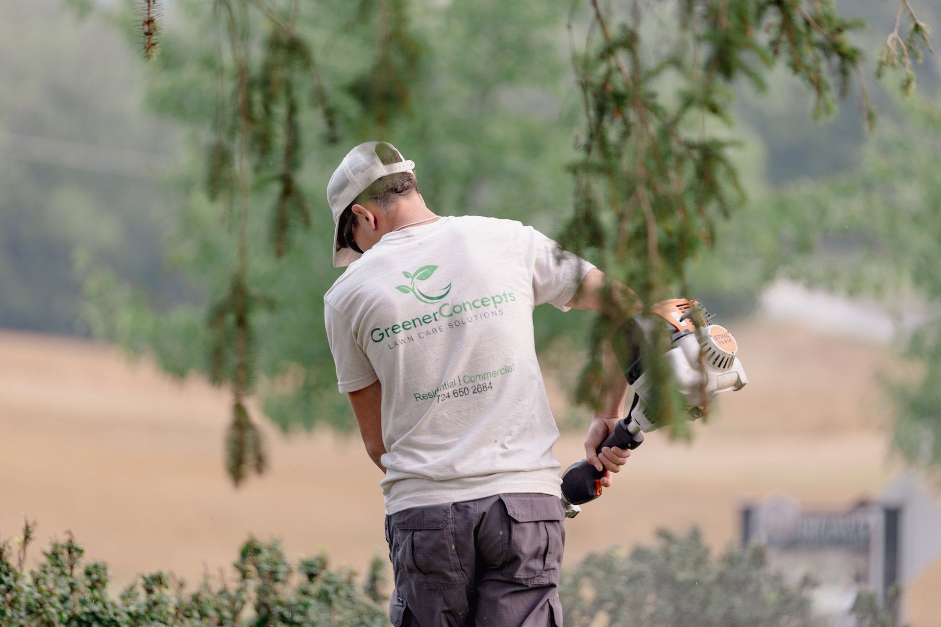 Person carrying a fishing rod and a fish by a lakeside, wearing a cap and gray shirt.