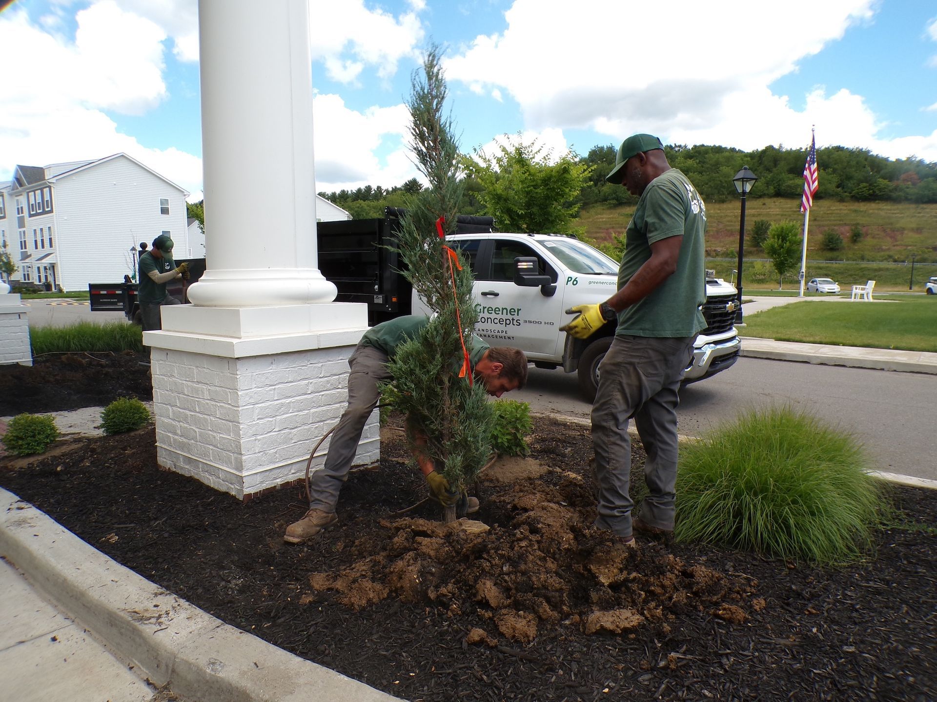 Man planting shrubs in a landscaped flower bed beside a white column and parked truck