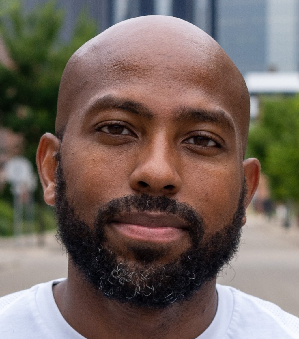 Man with a shaved head and beard, smiling, outdoors.
