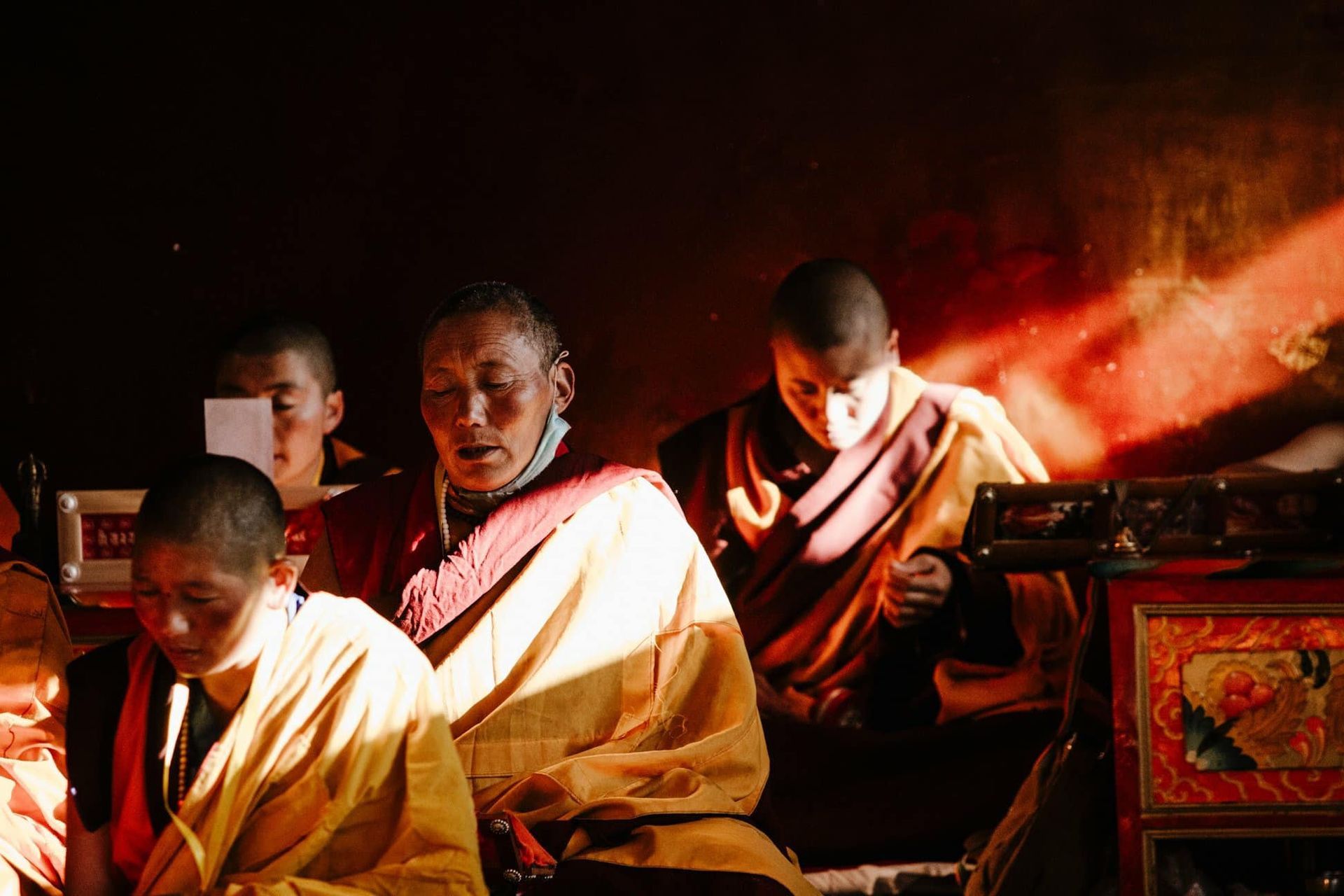 Monks in red and gold robes chanting in warm candlelight inside a dim temple