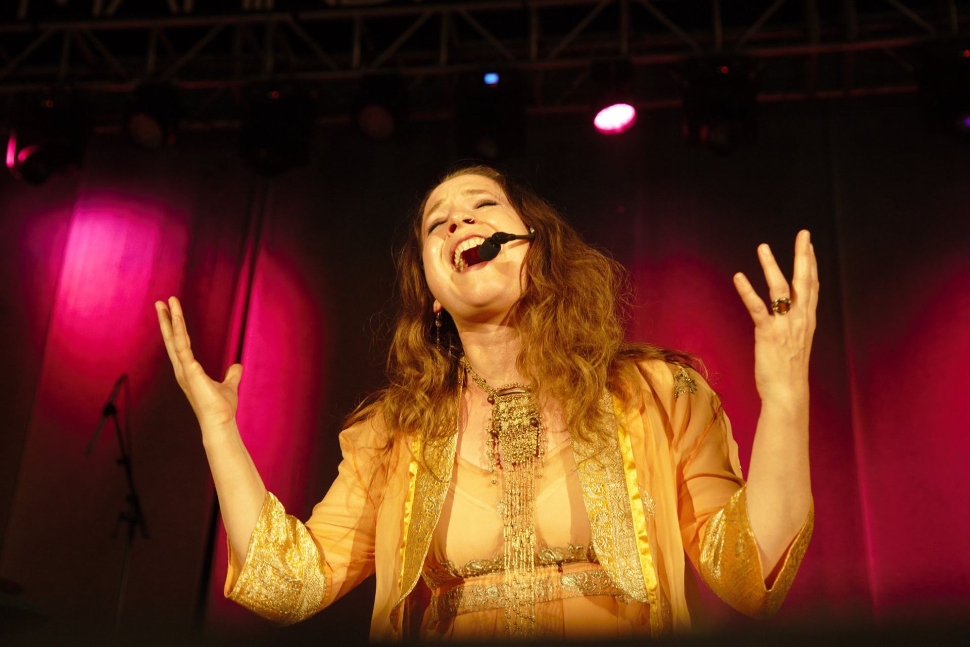 Woman singing on stage with arms raised, lit by pink and purple lights.