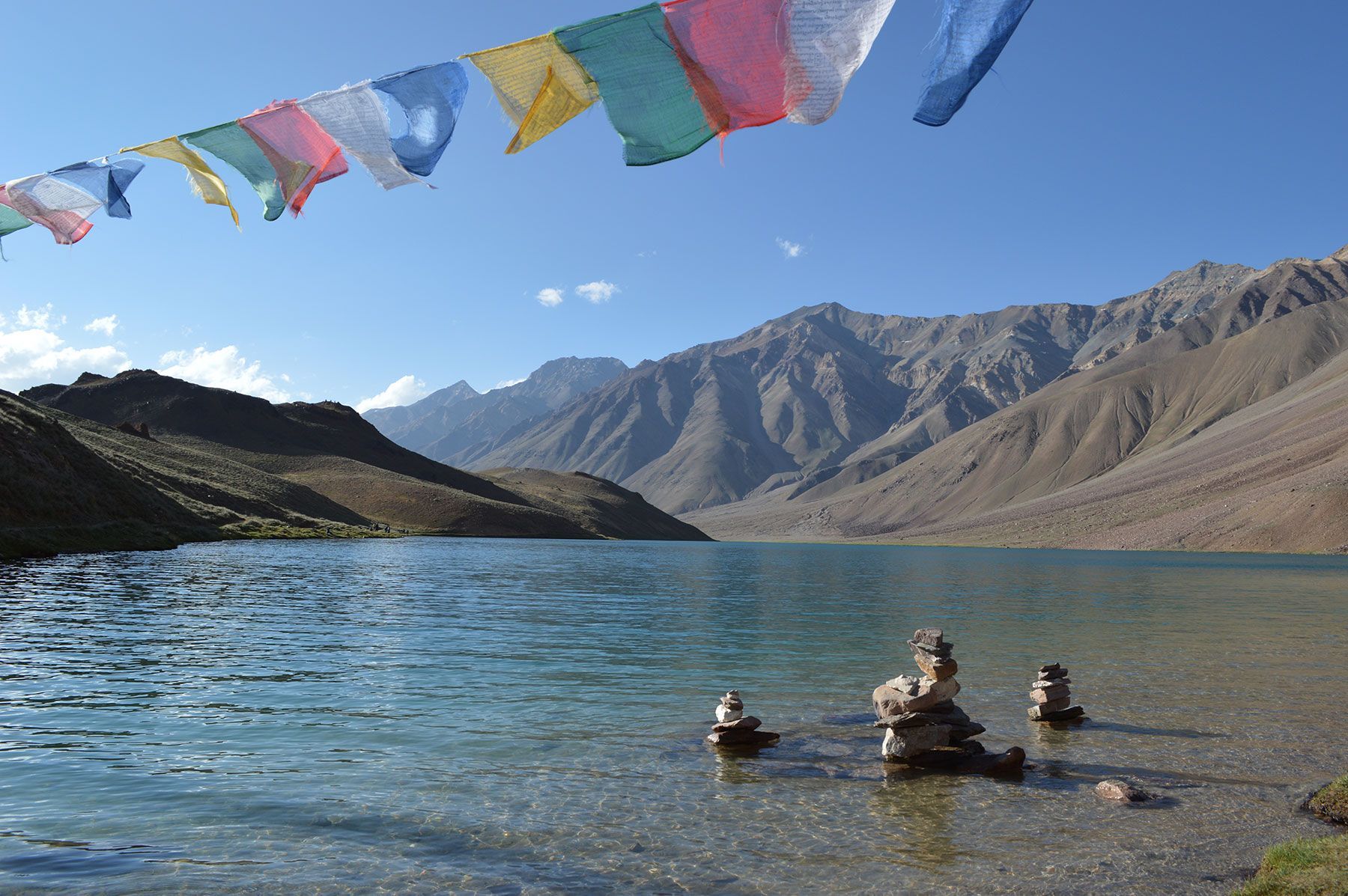 People wading in a turquoise lake with colorful prayer flags and mountains under a clear blue sky
