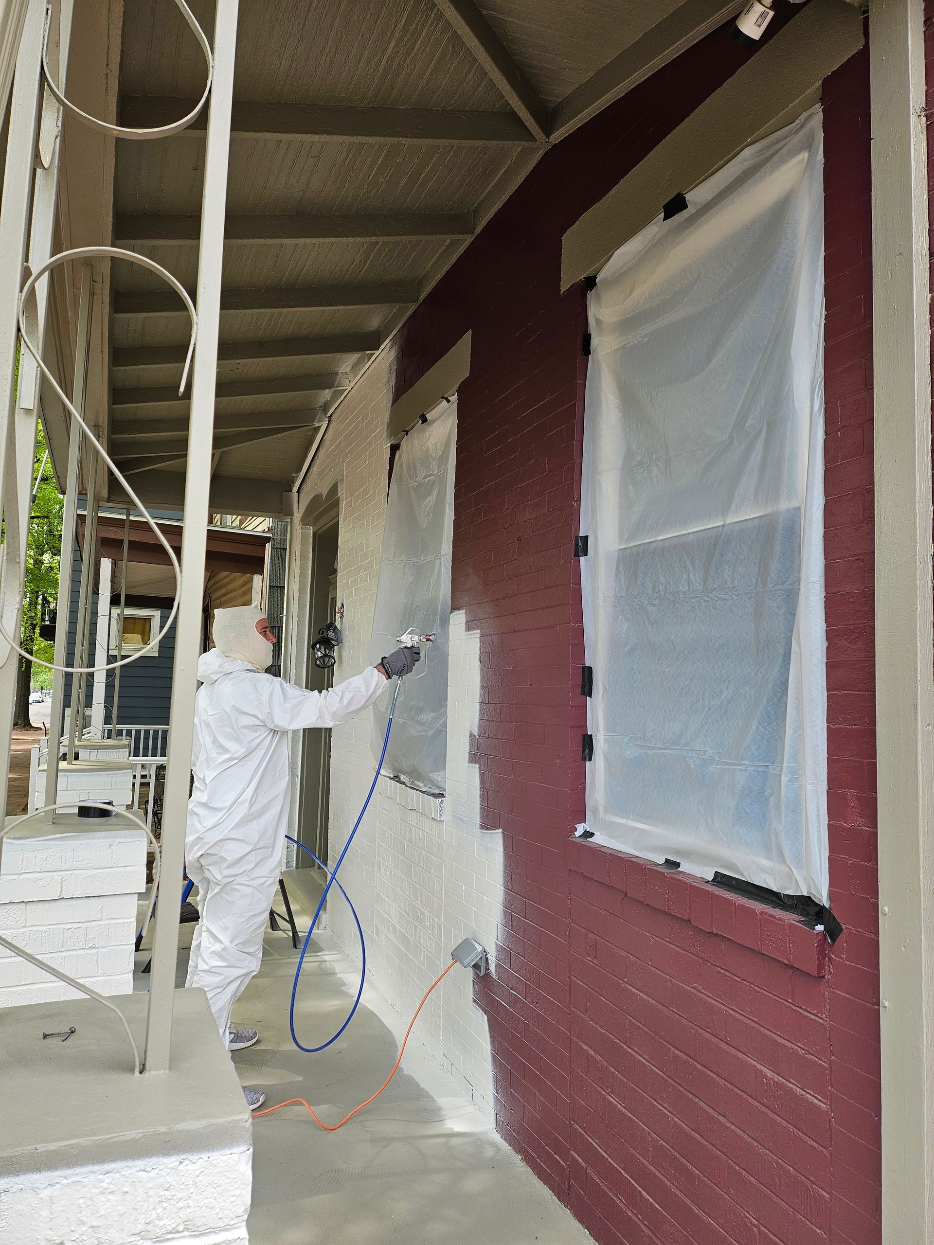 Person in white coveralls spray-paints a maroon building exterior under a covered walkway.