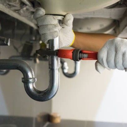 Gloved hands tightening a pipe under a sink with a red adjustable wrench