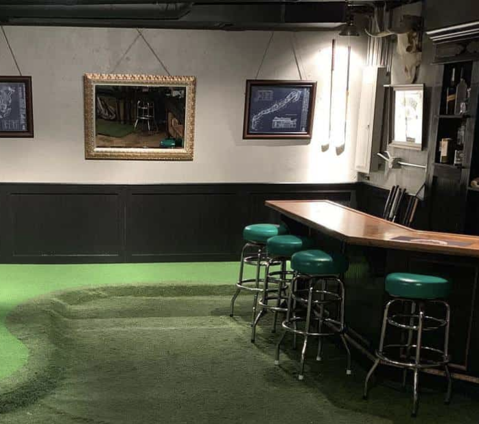 Dimly lit indoor bar with green carpet, wooden counter, and green-topped stools along the right wall